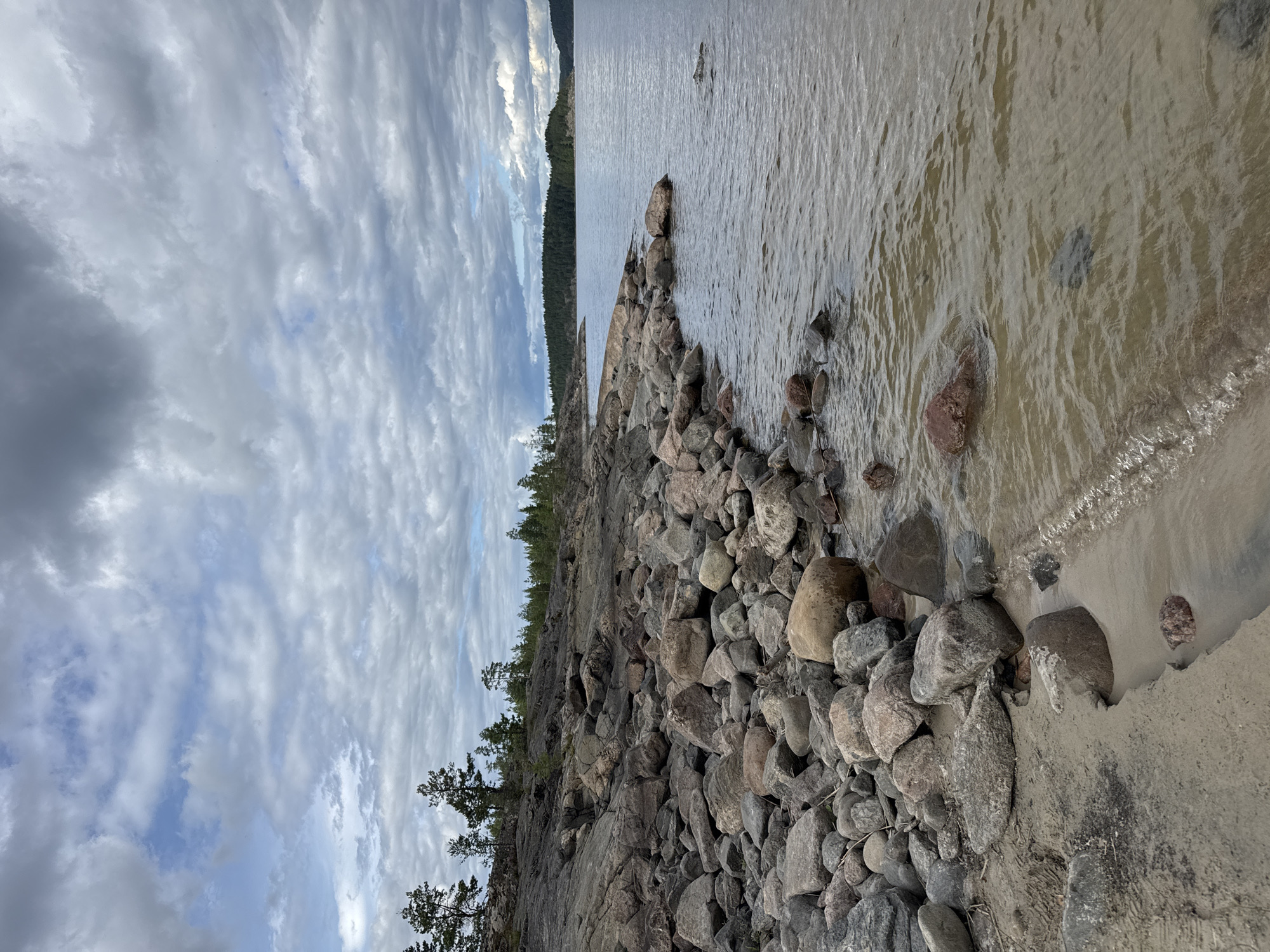 The image depicts a rocky shoreline along a body of water, likely a lake or bay, under a cloudy sky. The foreground is dominated by a beach of sand and an abundance of stones and boulders of various sizes and colors, ranging from light grey to reddish-brown. Some of these rocks are partially submerged in the shallow, murky water. The water itself is calm with gentle ripples.
Beyond the immediate shore, the landscape rises into a rocky, uneven terrain sparsely populated with coniferous trees. Further in the distance, a dense forest of evergreen trees covers the rolling hills of the opposite shoreline. The sky is filled with heavy, grey clouds, with hints of blue sky peeking through in some areas. The overall impression is of a natural, rugged, and somewhat overcast environment.
Based on the provided context, this scene is located in the city of Sorola, Russia, known for its numerous lakes and forested landscapes.