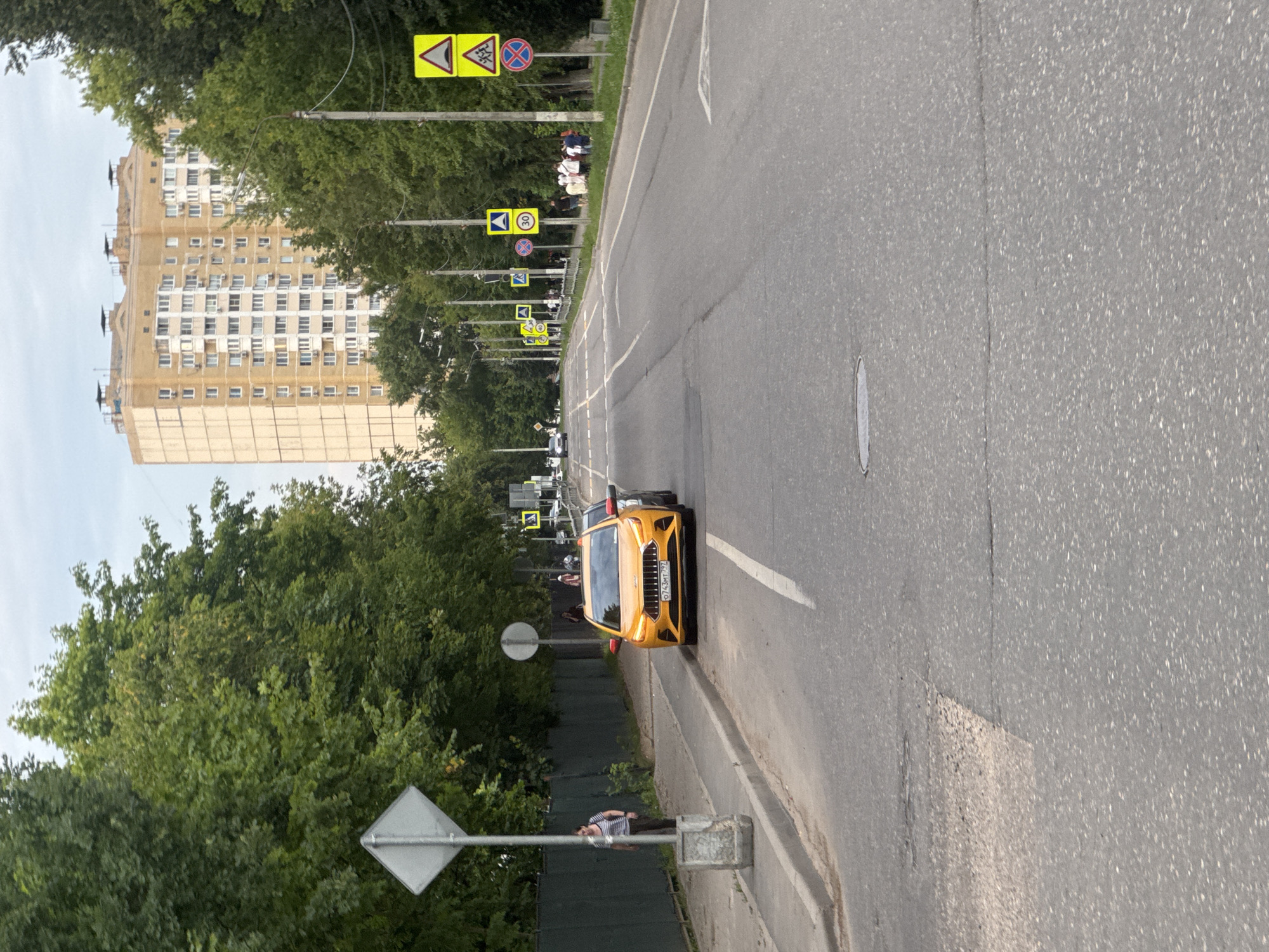 The image shows a street scene in Dedovsk, Russia. The foreground features a light yellow car parked on the right side of the road, facing away from the camera. The car has the license plate "O743MT 797". To the left of the car, a woman in a striped shirt and dark pants stands on the sidewalk, facing the street. Behind her, a diamond-shaped sign is visible. The road itself is paved and shows some wear and tear, with a manhole cover in the lower center.
The middle ground of the image displays a street lined with trees on both sides. Several traffic signs are posted along the right side of the road, including a speed limit sign (30), pedestrian crossing signs, and a general warning sign. A tall, light-colored apartment building is visible in the background, towering over the trees. A group of people are gathered on the right sidewalk, some appearing to be conversing.
The background is dominated by a cloudy sky and more trees. The overall impression is a suburban street on a cloudy day.