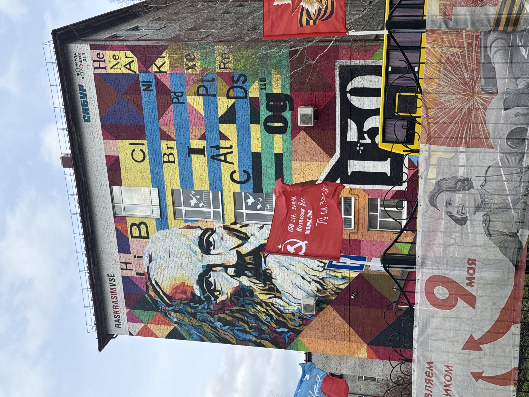 The image depicts a colorful mural on the side of a building in Tobolsk, Russia. The mural includes the periodic table of elements, a portrait of a person (possibly a historical figure), and the word "SIBUR," a Russian petrochemicals company. Text on the building also reads "RASKRASIMVSË" which means "We paint everything" in Russian. Red banners with Soviet-era text and symbols hang from the building. A banner with the number "9" and the word "мая" (May in Russian) is attached to a metal fence in the foreground, likely referring to Victory Day.