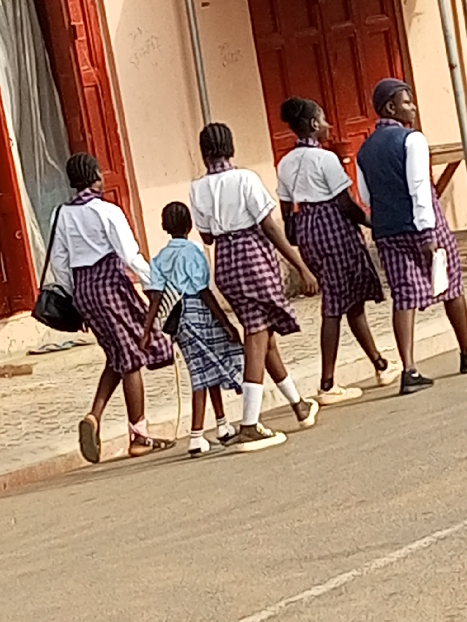 In Tumfure, Nigeria, five young girls in school uniforms are walking on a paved road in front of a building with red doors. The girls are wearing plaid skirts and white tops, and some have vests. The girl on the far left is carrying a black bag. The girl second from the right is holding a white object. The girls are walking away from the camera. The building appears to be a commercial establishment or residence, with a light-colored facade and a few windows visible. The road is textured and shows signs of wear. The lighting suggests it is daytime, possibly mid-morning or afternoon. The scene is captured from a slightly elevated angle.