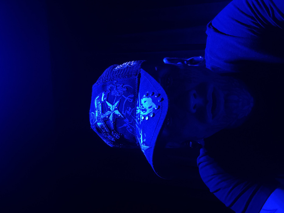 A wrestling mask adorned with a star and a skull sits on a person's face. The person is turned sideways, with their chin tilted down, and they are wearing a long-sleeved shirt. The entire scene is bathed in deep blue light, obscuring details and creating strong shadows. The mask has intricate designs and embellishments.