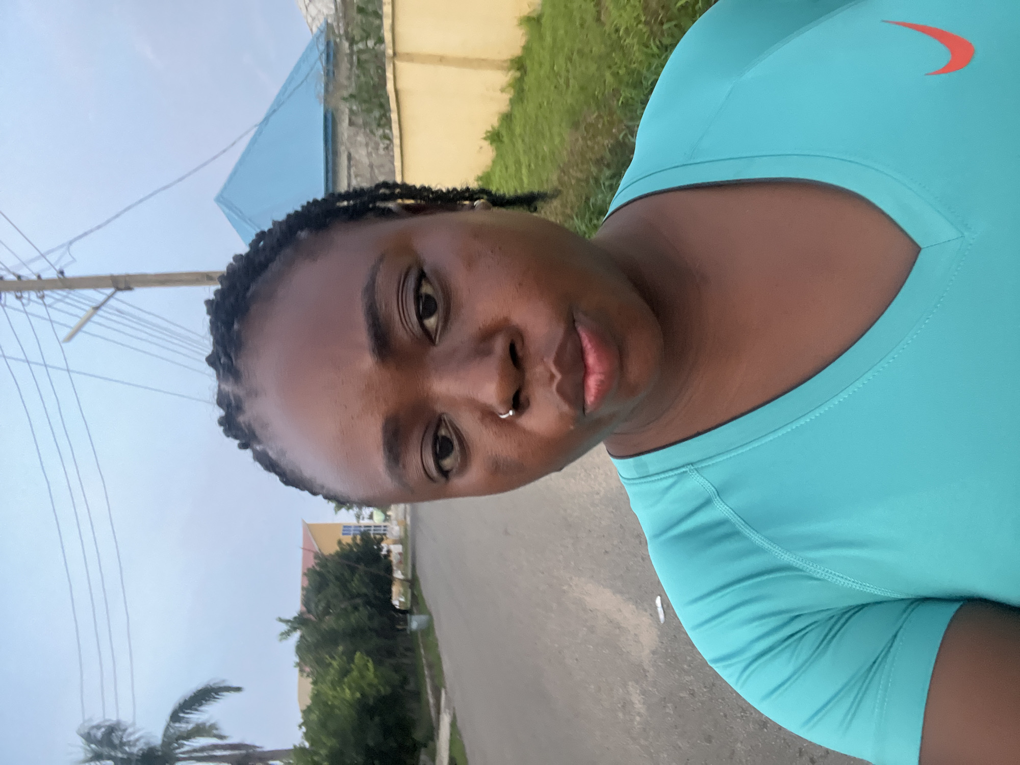 A medium shot captures a woman of color from the chest up, facing the camera in a street scene in Abuja, Nigeria. She has dark skin, black braided hair pulled back, and is wearing a bright turquoise V-neck t-shirt with a red Nike logo on the right side of her chest. A silver nose ring adorns her left nostril. Her expression is neutral, with her lips slightly pursed.
The background depicts a paved road, suggesting a residential or suburban area. On the left side of the road, a green palm tree stands tall, followed by dense green foliage and a yellow building with blue window frames. A trash can is visible near the side of the road. To the right of the woman, a tall yellow wall runs along the property line, with lush green grass and bushes behind it. In the distance, a house with a distinctive bright blue, pyramidal roof is visible, along with a utility pole carrying multiple wires across the sky. The overall lighting indicates an overcast day, typical of Abuja's climate.