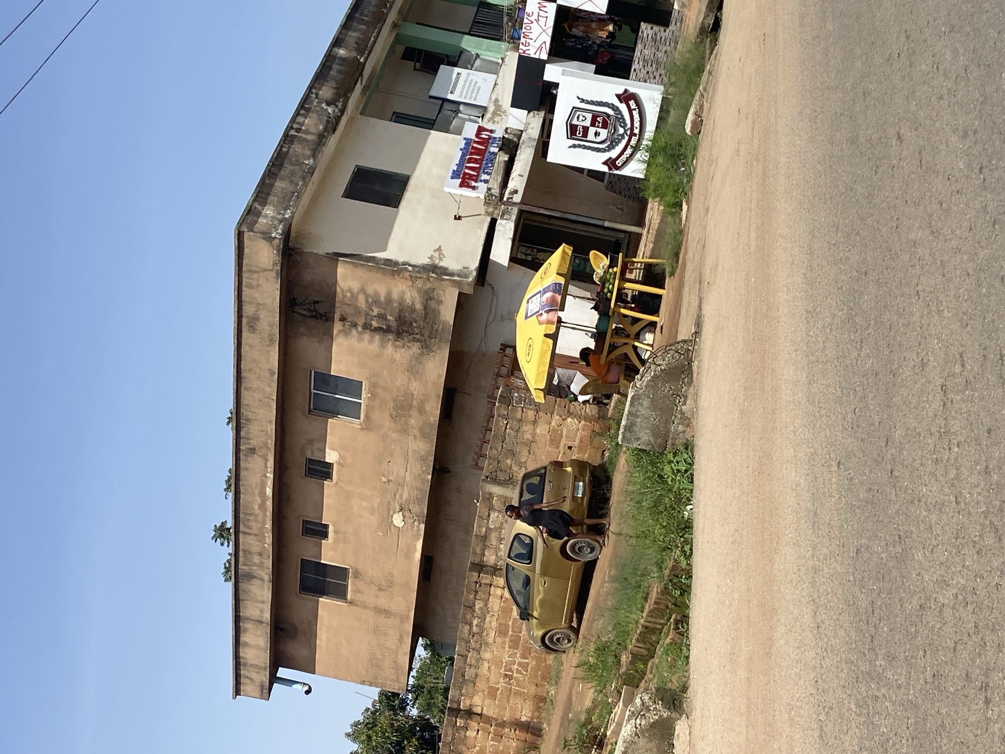 The image shows a street scene in Jos, Nigeria, with a focus on a two-story building. The building is a mix of old and new construction, with a weathered brown section on the left and a lighter colored section on the right. The right side of the building has a sign that reads "Watershed PHARMACY & STORES LTD." Another sign below it displays the logo for "CITYPOINT INT'L ACADEMY, JOS." A yellow umbrella is set up outside the pharmacy, providing shade for a table with some fruit. A woman in a black dress stands next to a small golden car parked on the dirt road in front of a brick wall. The road is unpaved and dusty, with some overgrown vegetation. The sky is clear and blue. The scene depicts a typical commercial and residential area in the city.