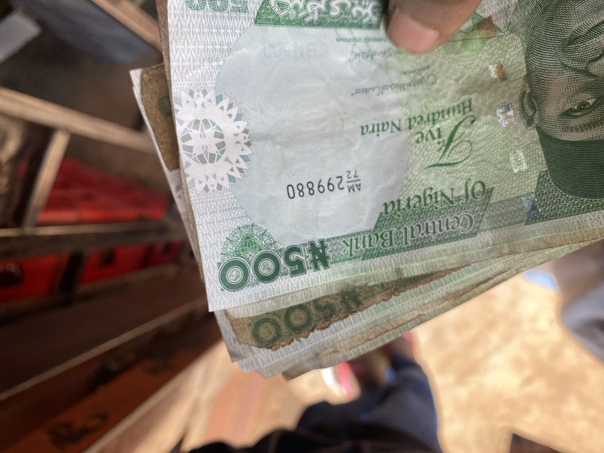 This media file is a close-up shot of several Nigerian 500 Naira banknotes being held by a person's hand. The banknotes are predominantly green and feature intricate patterns and the portrait of a man. Text visible on the banknotes includes "Central Bank Of Nigeria," "Five Hundred Naira," and a serial number, "AM 72 299880." The banknotes appear to be slightly worn, with some creasing and smudging, suggesting they have been in circulation. The background is blurred, but it seems to be an indoor setting with some wooden or metal structures and hints of red and brown colors. The lighting suggests it could be daytime, but the specific time or weather conditions are not discernible from the image. The focus is entirely on the currency and the hand holding it, with no clear indication of the specific activity or event beyond the handling of money. The image is taken from a slightly downward angle, emphasizing the denominations of the bills.