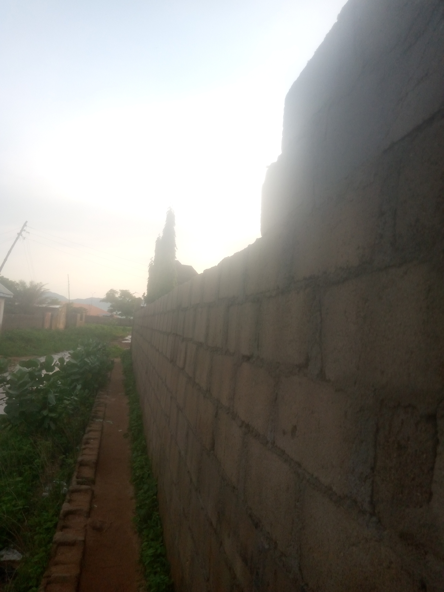The image captures a view of a brick wall constructed from concrete blocks, running diagonally from the foreground to the middle ground of the image, on the right side. The wall appears to be part of a residential or commercial property. To the left of the wall, there is a narrow dirt path bordered by vegetation. In the background, there are several buildings, some with corrugated iron roofs, and trees. The sky is bright and appears to be overcast or hazy, suggesting it might be early morning or late afternoon. In the far background, a faint outline of hills or mountains is visible. A utility pole with wires is also present on the left side of the image. The scene appears to be a street or pathway in a developing area of Jalingo, Nigeria, characterized by modest construction and a natural, somewhat unkempt environment. There are no people visible in the image.