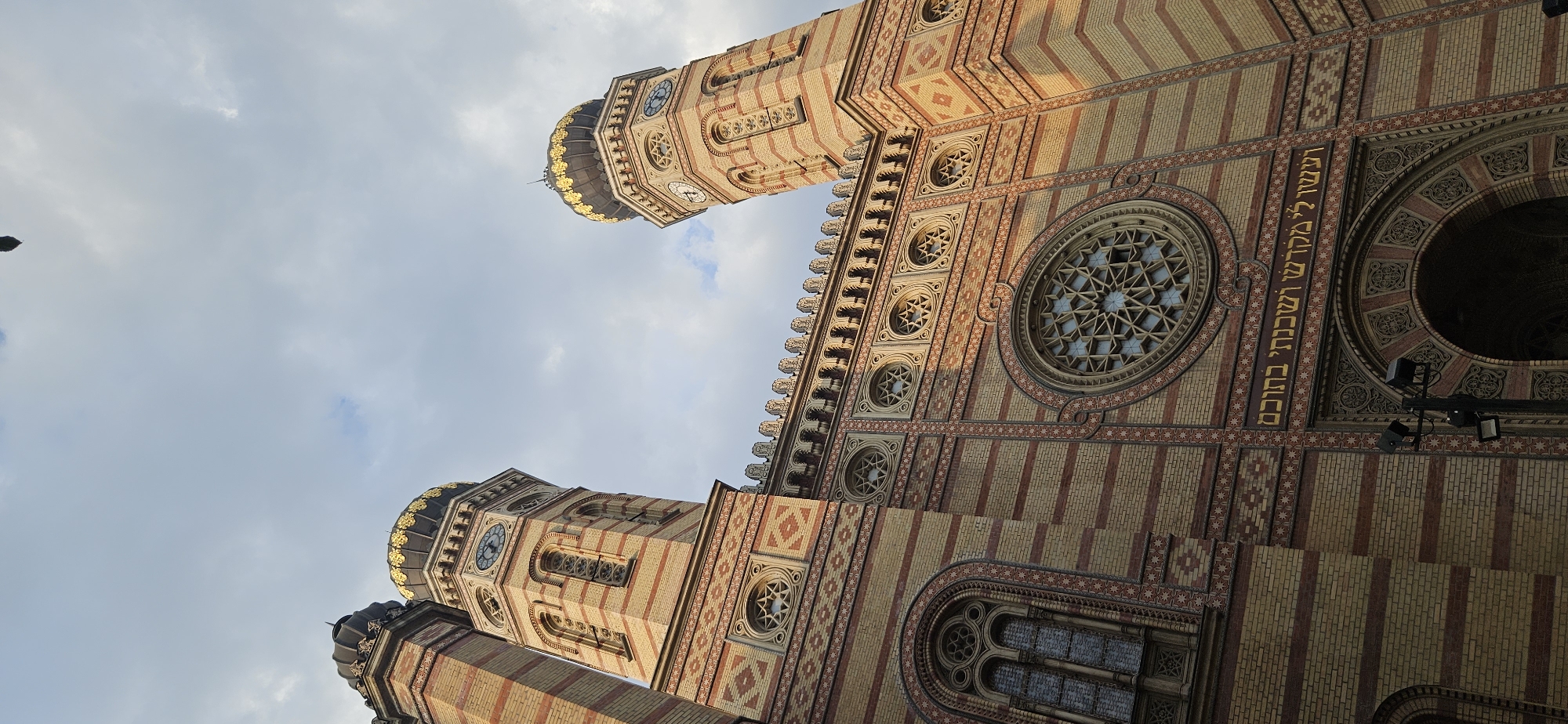 The image shows a low-angle, close-up view of the Dohány Street Synagogue in Budapest, Hungary. The ornate brick facade of the synagogue is visible, with its distinctive yellow and red striped pattern. Two towers with golden domes flank the central structure. The left tower has a clock face. In the center of the facade, above an arched entrance, there is a large rose window with intricate geometric detailing. A Hebrew inscription in gold lettering runs across the facade above the rose window. The sky above is partly cloudy. A lamppost with spotlights is visible in the lower right corner of the image. The overall impression is one of grandeur and architectural detail.