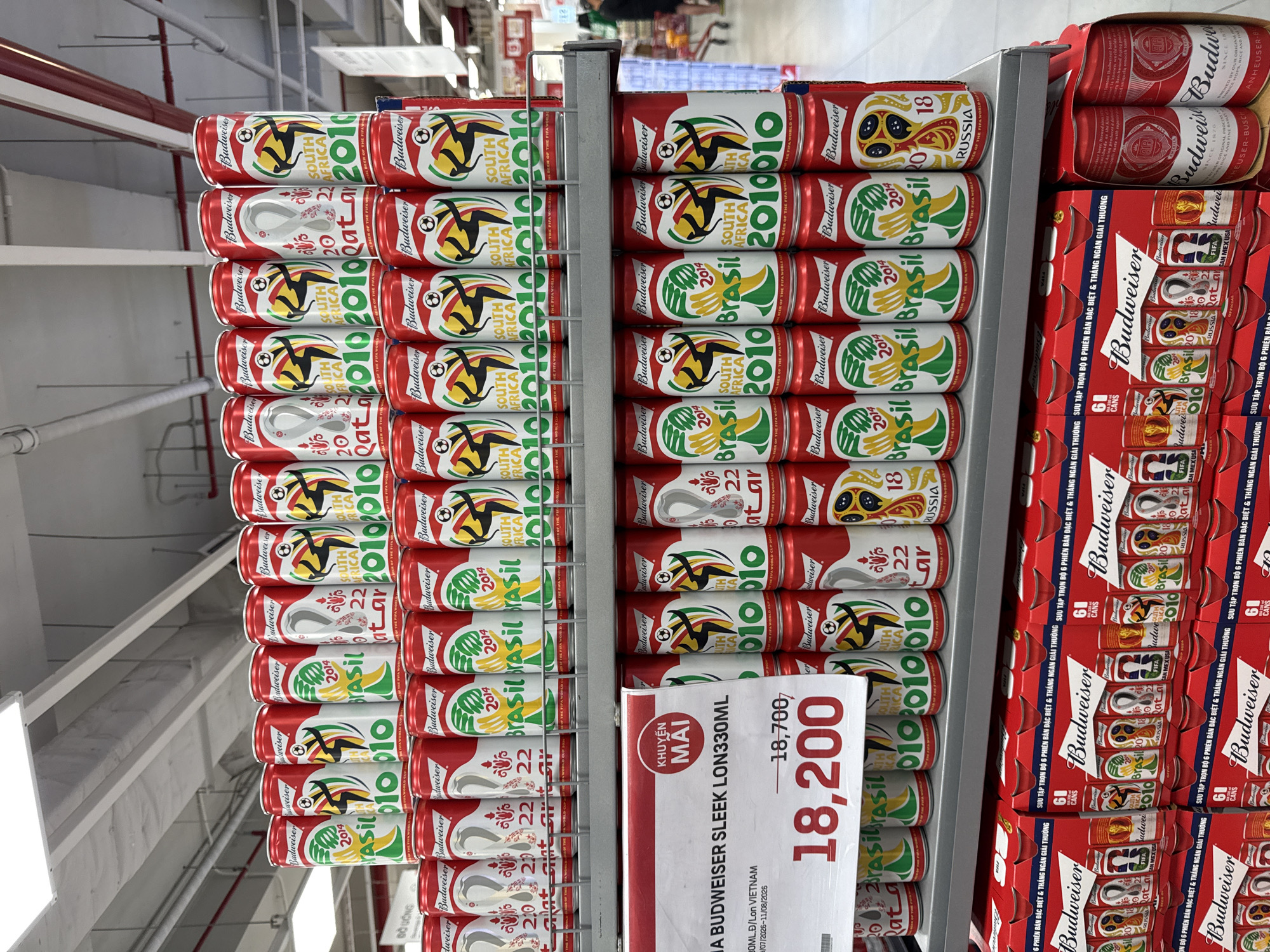 The image shows a supermarket shelf in Nha Trang, Vietnam, displaying multiple rows of Budweiser beer cans. The cans are red and feature designs commemorating various FIFA World Cups, including designs for 2010 (South Africa), 2014 (Brazil), and 2018 (Russia), as well as 2022 (Qatar). Some cans also have the Budweiser logo and "FIFA World Cup" text. Below the cans, there are displays of multi-packs of Budweiser beer in red boxes, also featuring World Cup-themed designs. A price tag is visible on the left side, indicating a price of 18,700 (presumably VND) with a sale price of 18,200. The environment appears to be an indoor retail space with fluorescent lighting and shelving units. No people are clearly visible in the immediate foreground or interacting with the products. The overall scene depicts a commercial display of alcoholic beverages in a retail setting.
