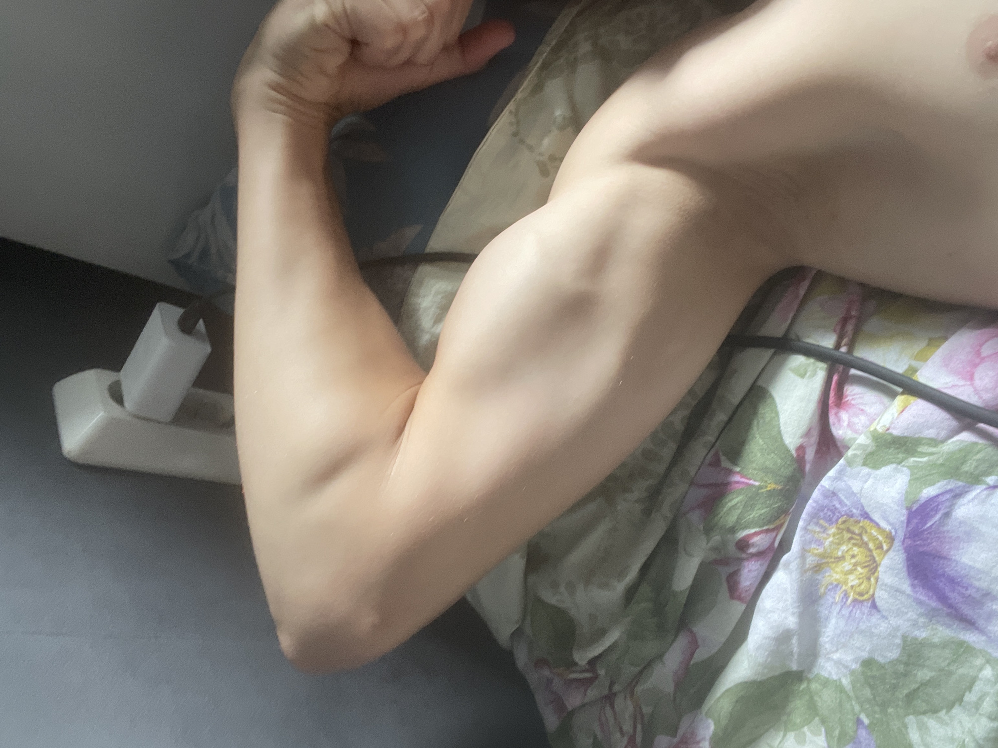 A close-up, top-down shot shows the arm of an individual lying on a floral-patterned surface. The arm is bent at the elbow, with the bicep clearly flexed. The skin appears light-toned and smooth. A white electrical outlet is visible in the upper left corner of the frame, partially obscured by the arm. The background consists of a dark, textured surface, possibly a wall or bedding. The lighting suggests an indoor setting with light coming from the right. The scene is devoid of any discernible urban or city elements, making it impossible to determine a location within a city.