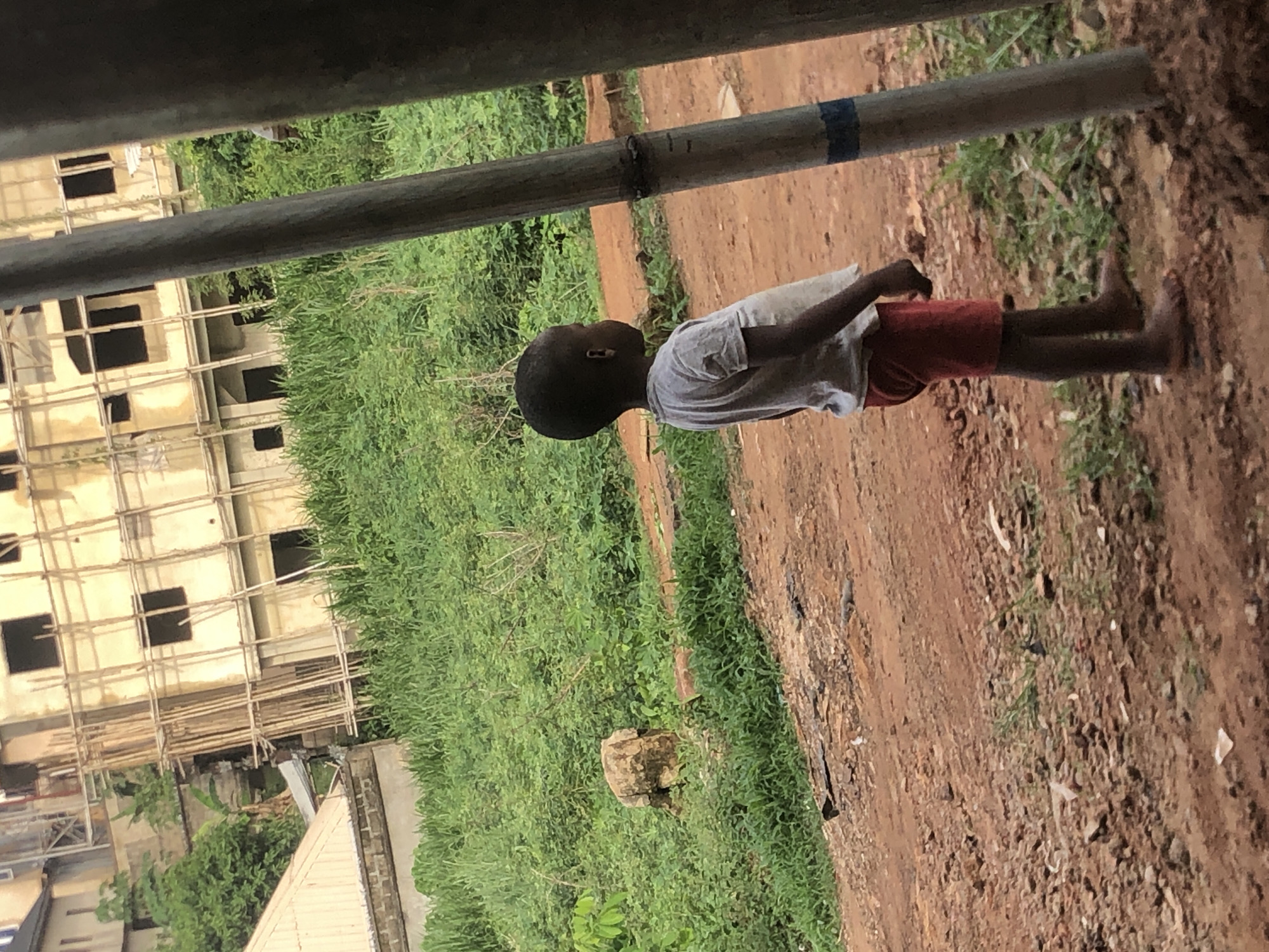 The scene is located in Nkwelle Ezunaka, Nigeria. The photograph depicts a young child standing on a dirt path beside a grassy area. The child is wearing a light-colored shirt and reddish-orange shorts and is barefoot. Overhead, there is a metal pipe, appearing to be part of some infrastructure. In the background, there is a building under construction with scaffolding around it, partially obscured by vegetation. The dirt path is brown, and the vegetation consists of green plants and trees. The lighting appears overcast.