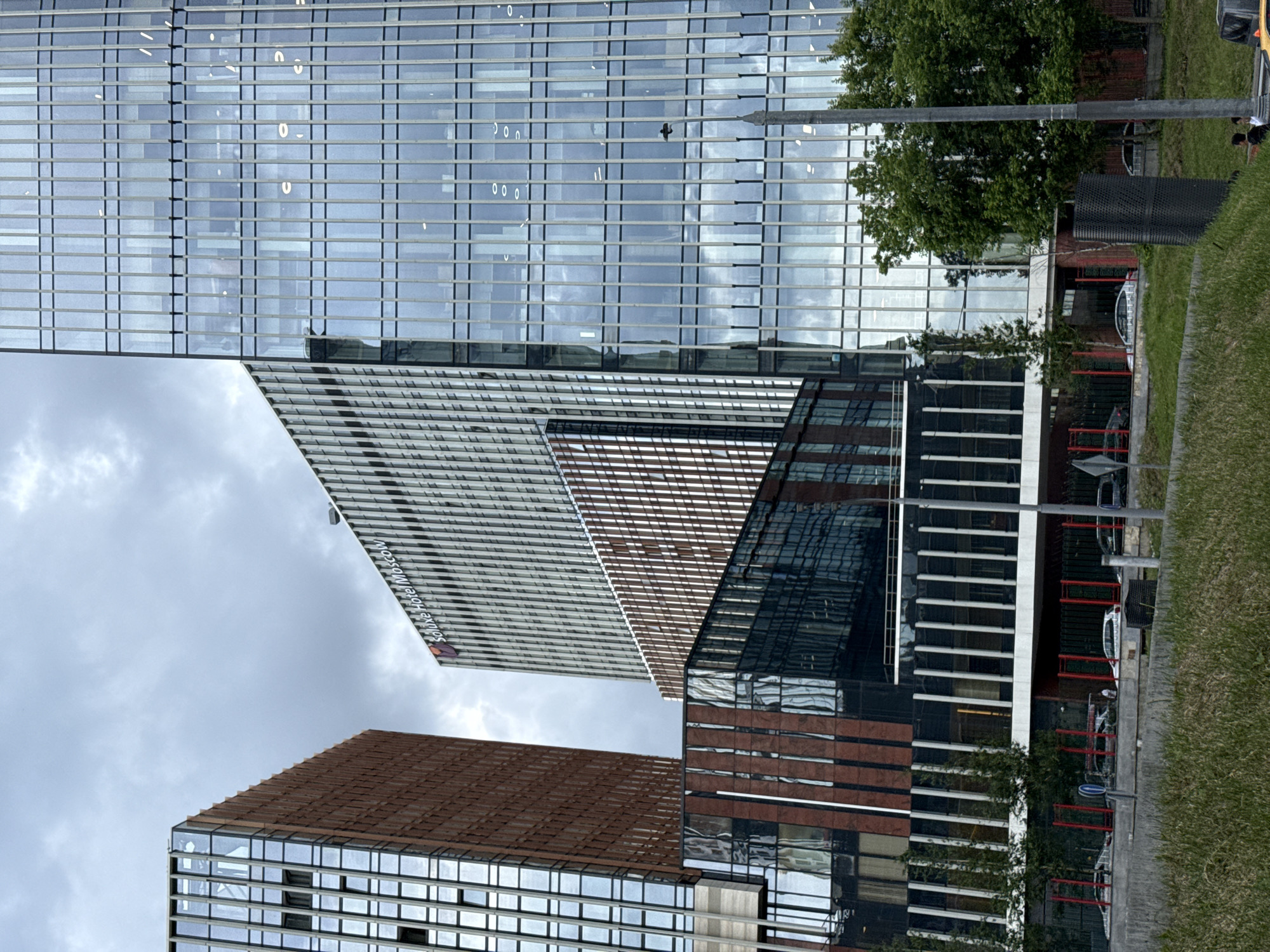 This image depicts the Soluxe Hotel Moscow, a modern building complex with a striking architectural design. The hotel is characterized by its glass facade, which reflects the surrounding environment and sky. Several buildings of varying heights and designs are visible, including a taller, more slender glass tower to the right, and a wider, partially brick-clad structure to the left. The lower portion of the complex features a dark, reflective surface with vertical red and white accents, suggesting a contemporary and sleek aesthetic.
In the foreground, a patch of green grass leads to a paved area with a low barrier and some parked cars. The sky is overcast with clouds, indicating a possibly cool or temperate day. The presence of a lamppost on the right side of the image suggests an urban setting. A couple of individuals are visible near the bottom right corner of the image, one standing and another in a seated position near a vehicle. The overall impression is of a well-maintained and modern urban environment, likely a business or hotel district within Moscow.