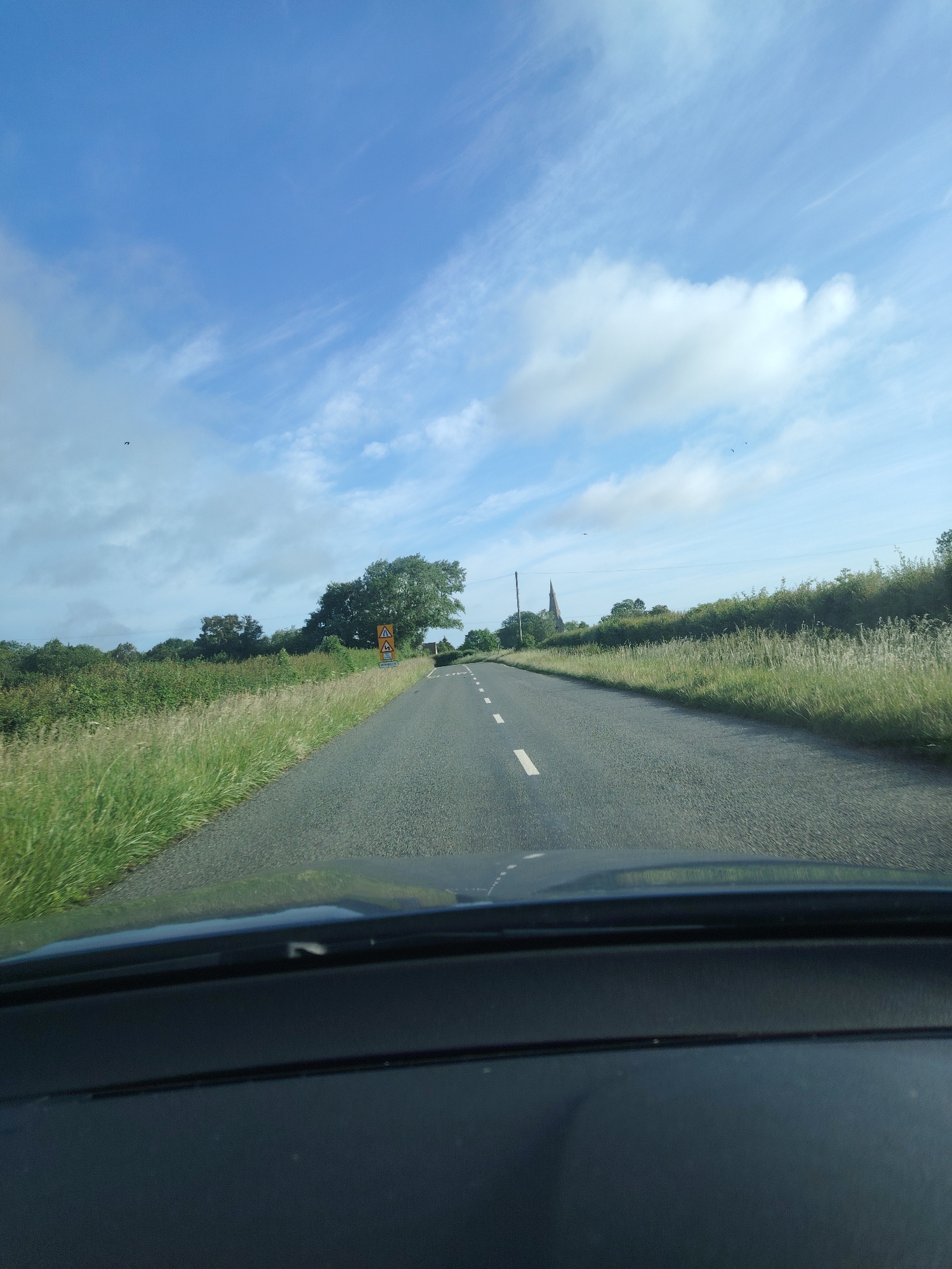 The image presents a forensic, matter-of-fact view from the interior of a vehicle traveling on a rural asphalt road in Huntingdonshire, United Kingdom, under bright daytime conditions. The sky is predominantly clear blue with scattered white cumulus and wispy cirrus clouds. Small dark specks, identified as birds, are visible against the sky in the upper left and upper right quadrants.
The road is dark grey asphalt, featuring a dashed white center line, indicating a two-lane configuration. It extends into the distance, gently curving towards the right. Both sides of the road are lined with wide grassy verges comprising tall, light green grasses. Beyond the verges, dense green bushes and trees form the landscape.
On the left side of the road, approximately mid-distance, a yellow diamond-shaped road sign depicting a car bouncing over humps (indicating an uneven road) is mounted on a pole. Below it, another smaller yellow diamond sign is visible, its specific symbol less discernible but consistent with UK road signage. Further down this side, a line of dense, dark green trees is visible against the horizon.
On the right side of the road, a similar grassy verge and a thick hedge line separate the road from further trees. In the far distance, a prominent, pointed church steeple or spire, constructed from light-colored masonry, rises above the tree line. A wooden utility pole with visible lines stands on the right side of the road, positioned between the hedge and the distant church.
The foreground of the image shows the dark dashboard and the lower edge of the windshield of the vehicle from which the photograph was captured. No individuals or other vehicles are distinctly visible within the scene. The overall environment is rural and verdant, consistent with the geographical context of Huntingdonshire.