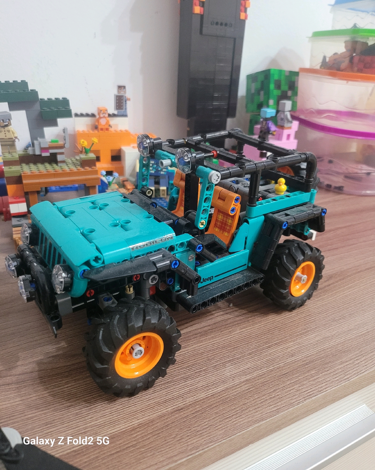 The image shows a close-up view of a teal and black Lego Technic Jeep Wrangler Rubicon model, prominently displayed on a wooden surface. The Jeep has orange-rimmed wheels with black tires. The interior of the Jeep features orange seats with a red plaid pattern and a small yellow rubber duck. The Jeep is positioned in the foreground, with a backdrop of various Lego structures. These include what appear to be Minecraft-themed Lego builds, such as a Creeper figure and a large black structure resembling a Nether portal or a figure from the game. There are also other Lego figures and structures in the background, including a brown wooden table or platform with some flowers. The lighting suggests an indoor environment. The image also contains a watermark in the lower left corner that reads "Galaxy Z Fold2 5G". No elements in the image suggest a specific location in Astana, Kazakhstan.