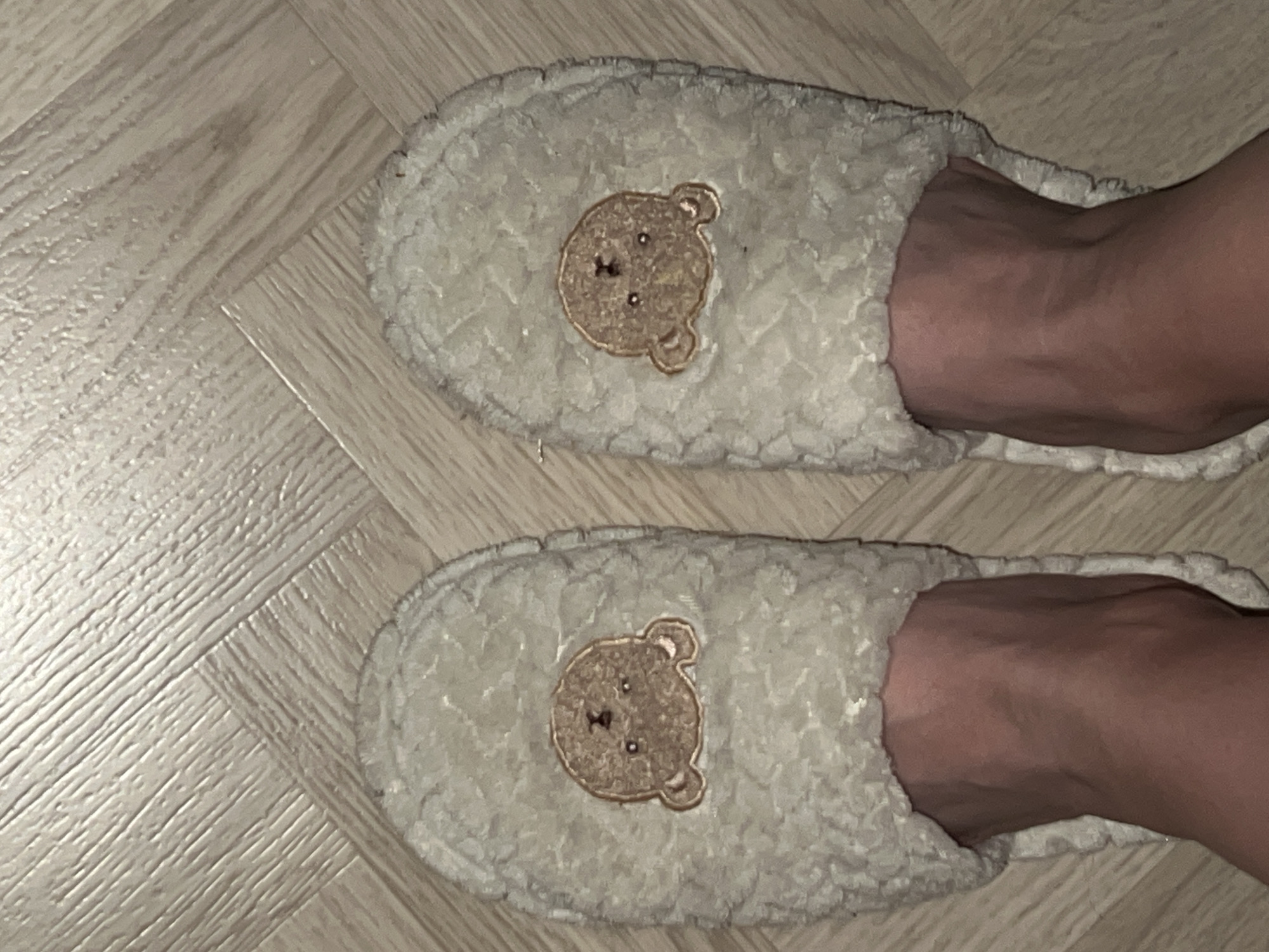 Two fluffy, cream-colored slippers with embroidered teddy bear faces are on a person's feet. The slippers are placed side-by-side on a light-colored wooden floor, which has a parquet pattern. The slippers are plush and appear to be worn indoors. The person's feet are visible emerging from the front of the slippers.