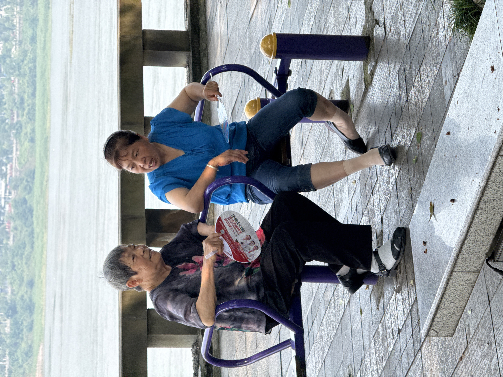 Two elderly Asian women are seated on purple outdoor exercise equipment on a wet, paved promenade in Jiangjin, China. The woman on the left, with short grey hair, wears a dark patterned short-sleeved top, black wide-legged pants, and black open-toe sandals over light grey socks; she holds a white and red promotional hand fan with Chinese characters, displaying a 2024 date and text related to "free access". The woman on the right, with dark hair pulled back, wears a blue short-sleeved blouse, dark blue cropped denim jeans, and black closed-toe flats over sheer, light-colored stockings; she has a silver-colored bracelet on her left wrist. Both women are facing each other, engaged in an animated conversation. Behind them, a low concrete railing separates the promenade from a wide, calm body of water, consistent with the Yangtze River. A distant green, tree-lined shoreline with visible structures is present across the water. The grey tiled ground beneath them is damp, with reflections and scattered small leaves. A grey stone bench or step is partially visible in the foreground.