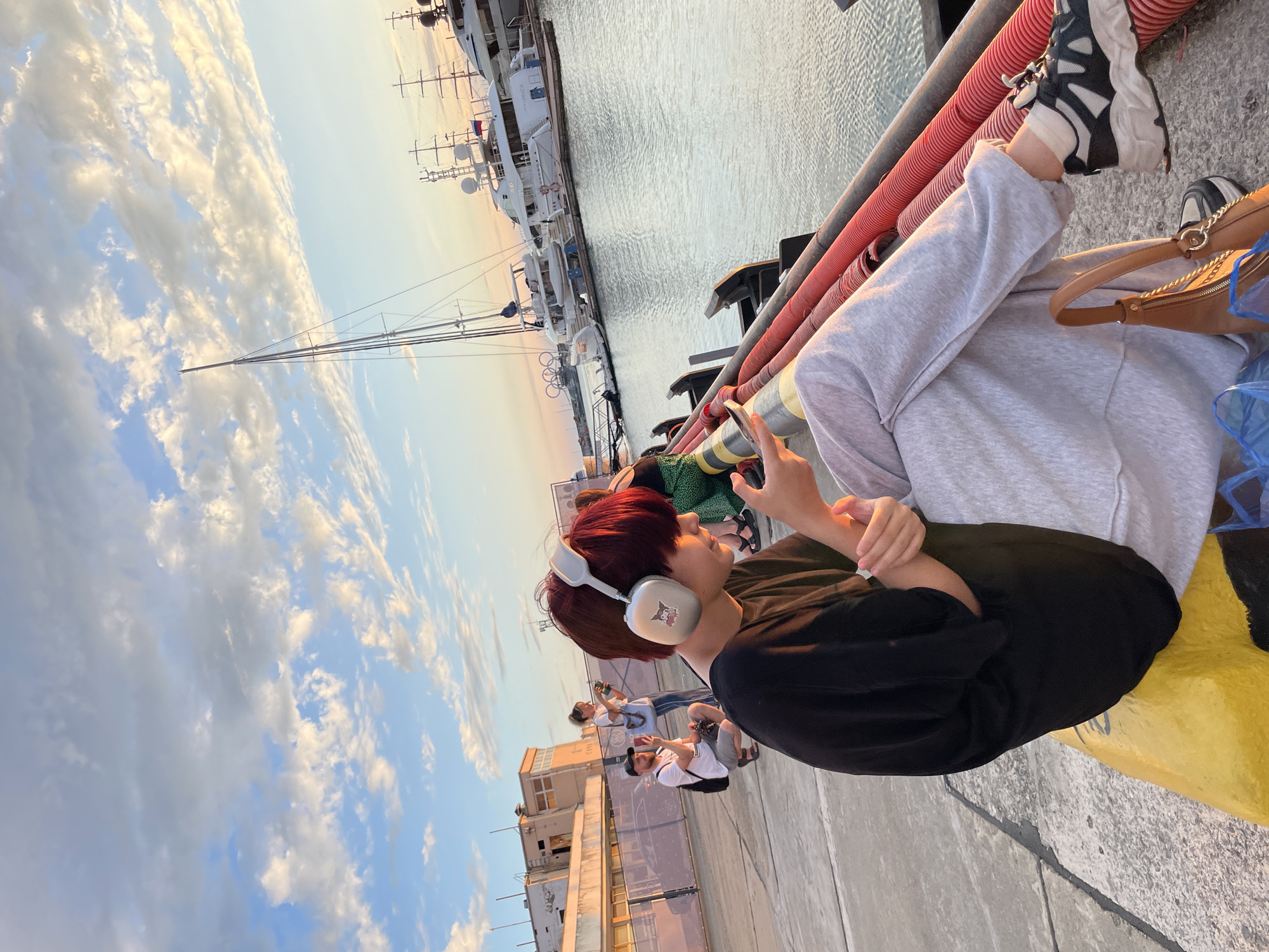 A group of people are gathered on a dock at sunset, enjoying the view of yachts and the sea. The sky is filled with dramatic clouds, bathed in the warm glow of the setting sun, suggesting a pleasant evening. The mood appears relaxed and casual, with some individuals listening to music with headphones, while others engage with their phones. One person, with striking red hair, is prominently featured in the foreground, wearing white headphones and a black t-shirt. Their posture suggests they are enjoying the moment and perhaps lost in thought or music. In the background, several large yachts are docked, indicating a likely affluent location. The presence of a small brown purse and the casual attire of the individuals suggest a leisurely outing. The concrete dock and metal railings are utilitarian, typical of a marina. The overall scene conveys a sense of tranquility and enjoyment of a beautiful evening by the water.