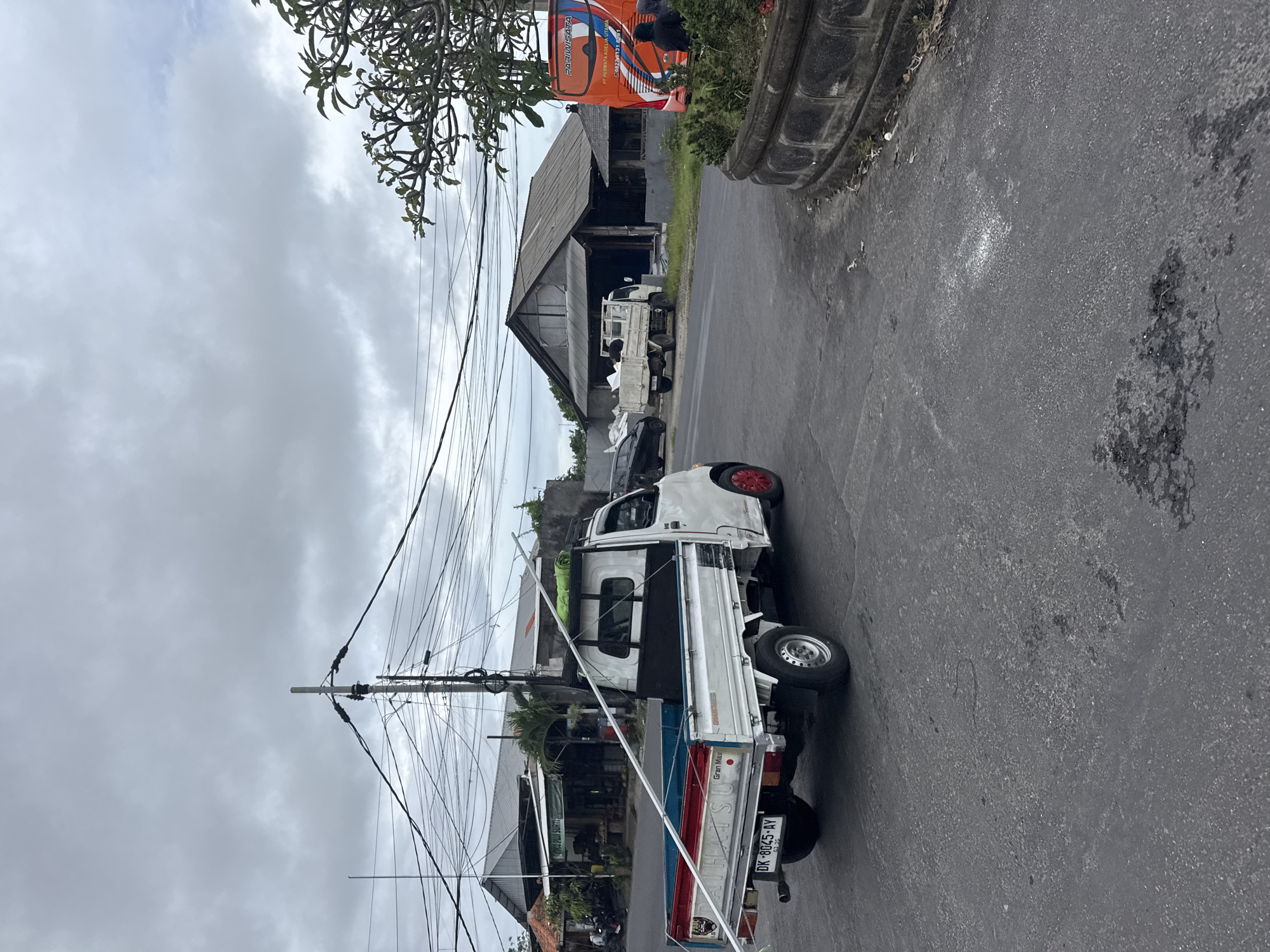 The image depicts a street scene in Kuta Selatan, Indonesia, under an overcast, cloudy sky. A white Daihatsu Gran Max pickup truck is prominently parked on the right side of an asphalt road, facing away from the viewer. Its tailgate features distinctive red and blue stripes, with the brand "Gran Max" and "DAIHATSU" visible, alongside the license plate "DK 8045 AY". The truck also has bright red front wheel accents.
To the left, a tall utility pole is heavily laden with numerous overhead electrical and communication wires that crisscross the frame. Beyond the pole, various buildings with sloped roofs are visible, interspersed with tropical foliage.
Further down the street on the right, a large orange bus is partially visible. Text on the bus includes "PURI WISATA", "P.O. PERMATA ABADI LOMBOK", and "SM.023-1133 BALI". Another smaller white pickup truck is parked behind the bus, near an older wooden building. The sky is entirely covered with grey clouds, indicating a dull or potentially rainy day. Tree branches with dark green leaves hang into the upper left corner of the frame. The road surface is dark asphalt, showing some lighter speckled areas and darker, possibly wet, patches. The scene suggests a regular day in a semi-urban or rural Indonesian setting, with no specific event or intense activity taking place.