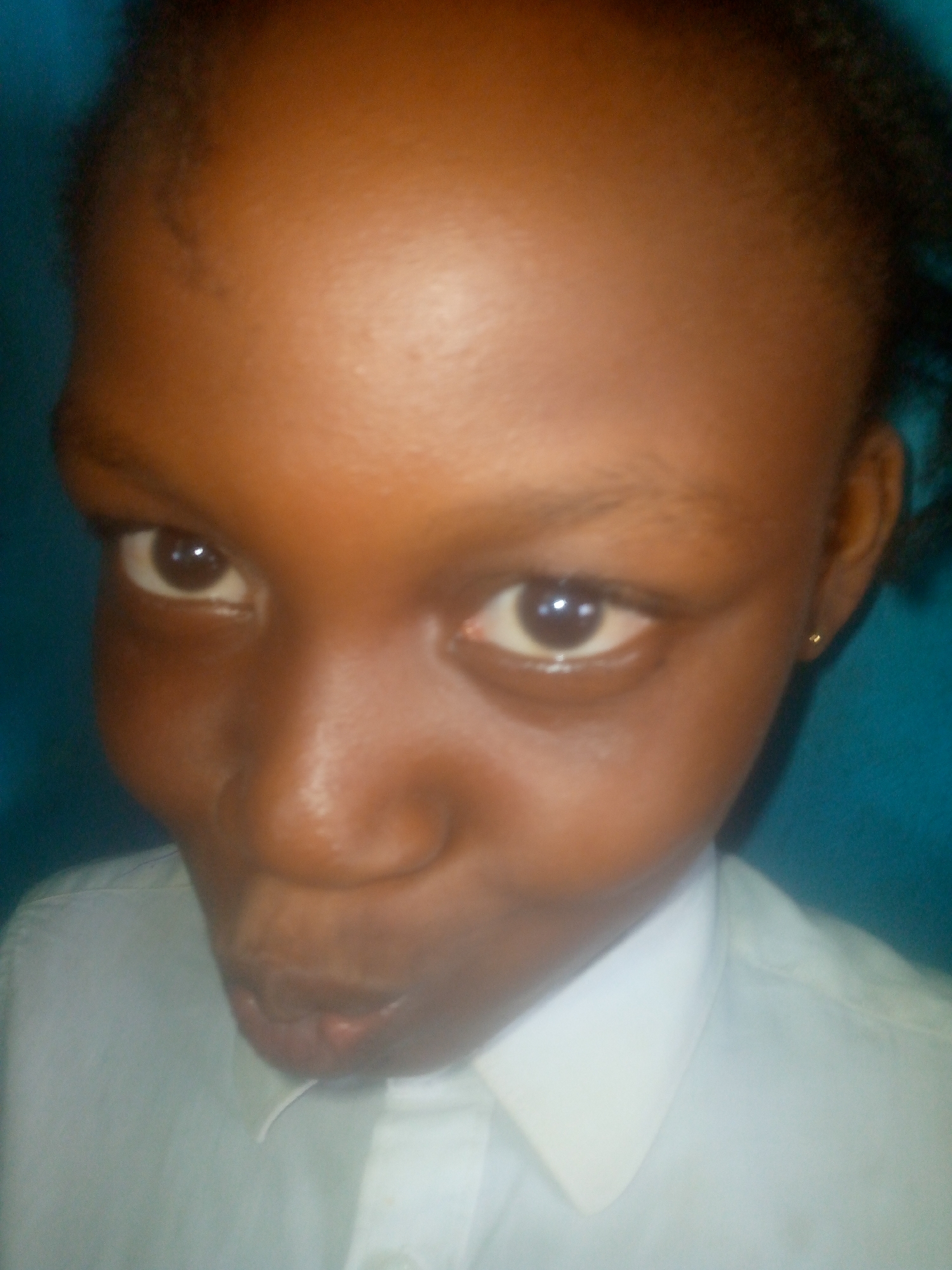 A close-up photograph depicts a young, dark-skinned individual with a smooth forehead, prominent eyes, and a slightly pursed mouth. They are wearing a white collared shirt. The location is unknown, and no further information is available from the image.