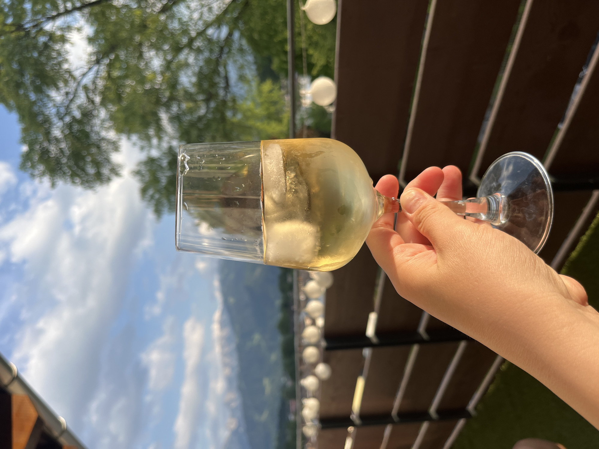 A hand holds a glass of white wine with ice, looking up towards a bright sky filled with fluffy white clouds. Trees with blurred green foliage are visible overhead, suggesting an outdoor setting. In the background, a row of white globe lights hangs above a wooden deck or pergola, and beyond that, a mountain range is faintly visible. The scene appears to be set in Garmisch-Partenkirchen, Germany, on a sunny day, likely in the afternoon given the lighting. The overall mood is relaxed and leisurely, possibly depicting a person enjoying a drink at an outdoor cafe or restaurant with a scenic view. There is no visible text in the image.
