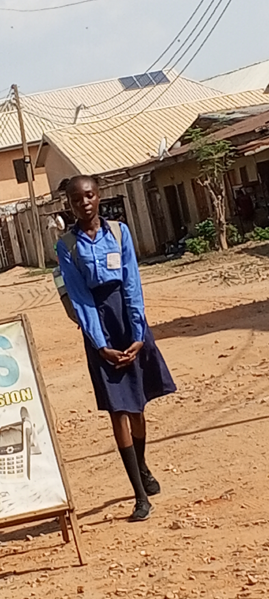 A young woman in a blue school uniform and black socks stands on a dirt road in Sabon Tasha, Nigeria. She has a backpack and is standing next to a sign. Buildings with corrugated metal roofs and power lines are in the background. One roof has solar panels.