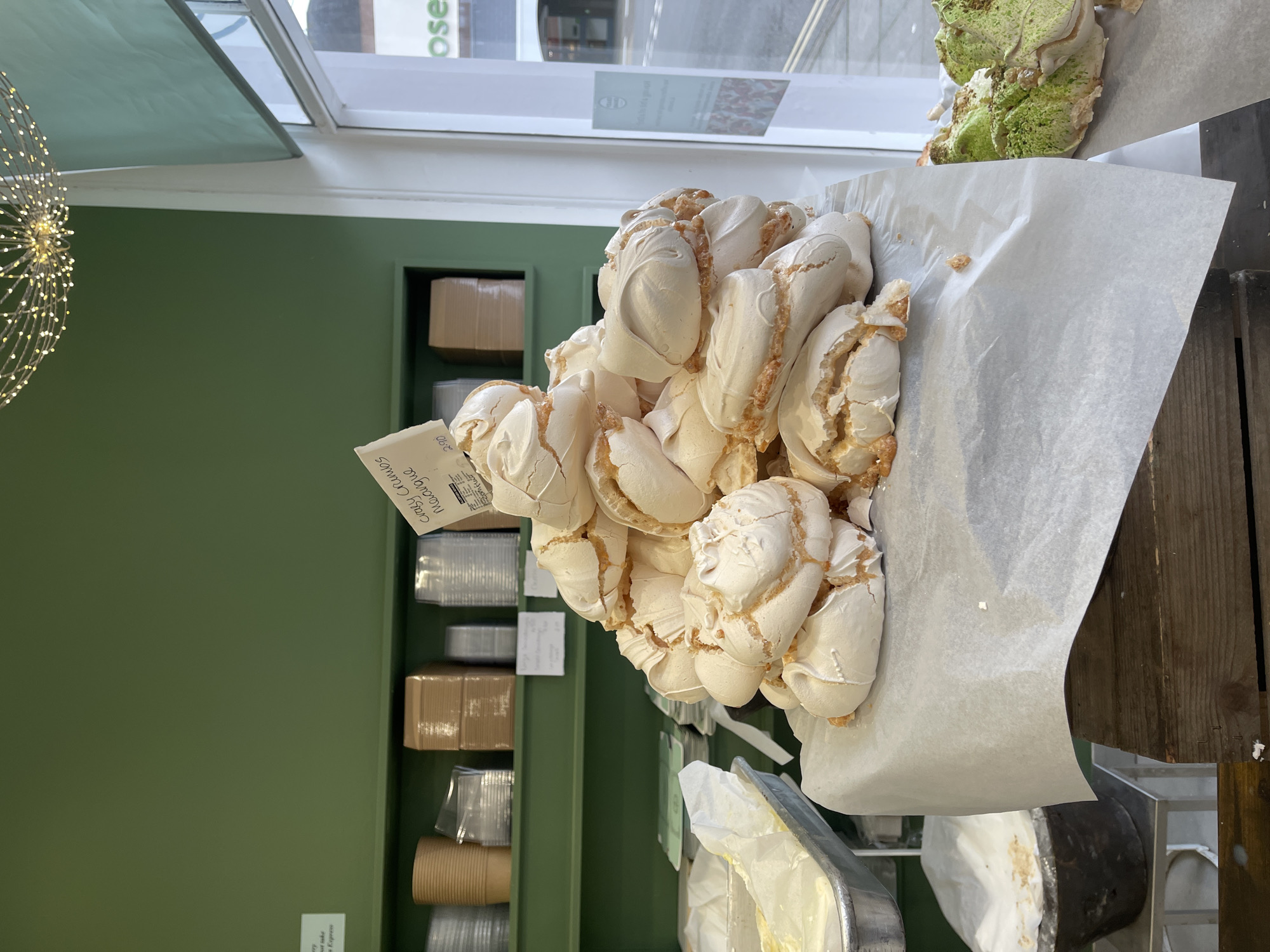 The image shows a pile of meringue cookies on parchment paper. A small price tag is visible, partially obscured by the meringue. In the background, there is a green wall with shelves containing disposable containers. The location is a cafe or bakery in Portsmouth, United Kingdom, as the address on the price tag is for that location.