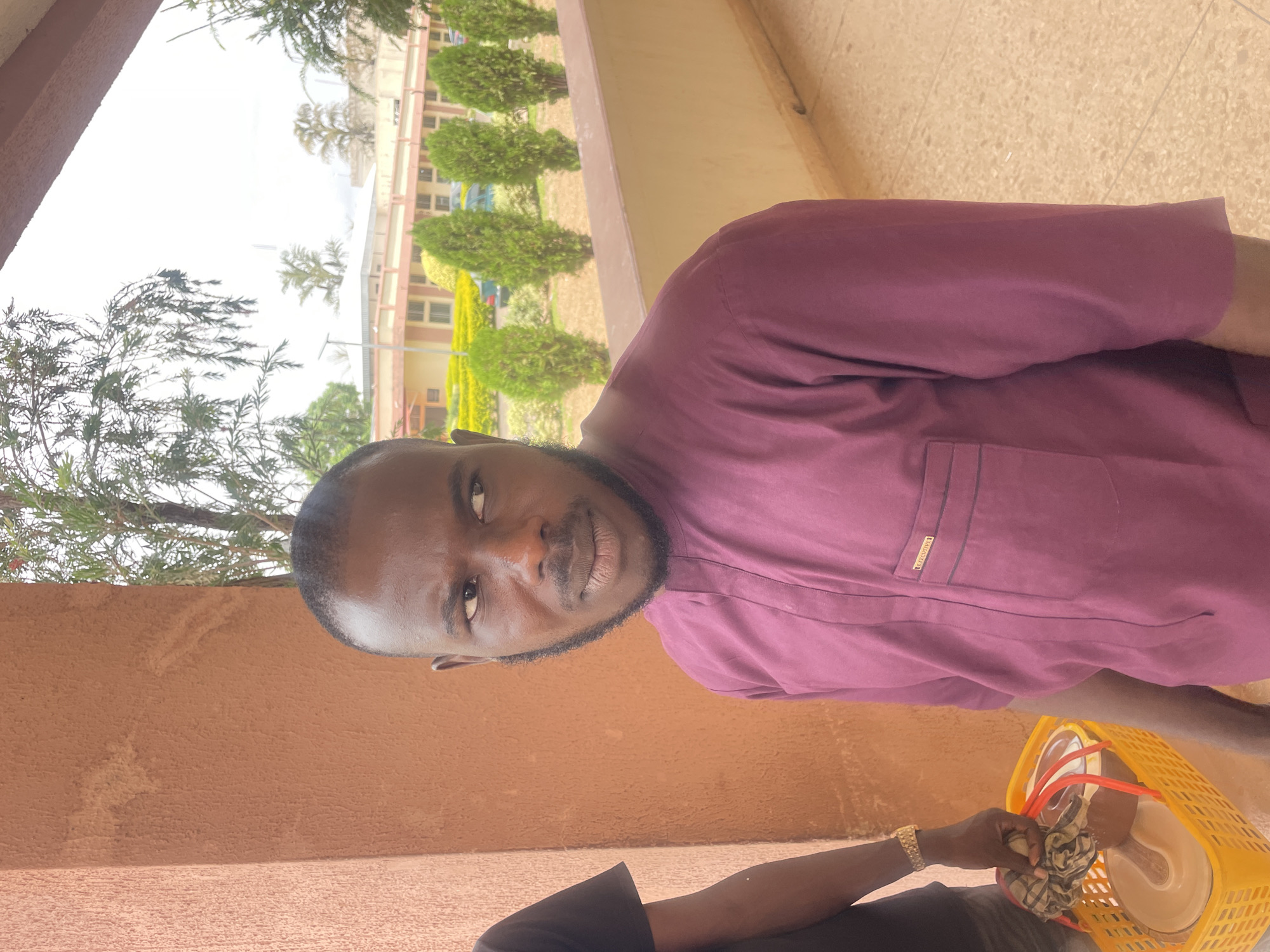 A medium-shot image depicts a man in Gwong, Nigeria, standing outside a building. He is wearing a maroon long-sleeved shirt with a pocket detail. He has short hair and a neatly trimmed beard. He holds a yellow plastic basket containing items including what appears to be plates wrapped in cloth. The background reveals the exterior of a multi-story building with multiple windows and landscaping featuring bushes and trees. A reddish-brown wall section appears in the foreground. The time of day appears to be daytime, evidenced by the natural lighting.