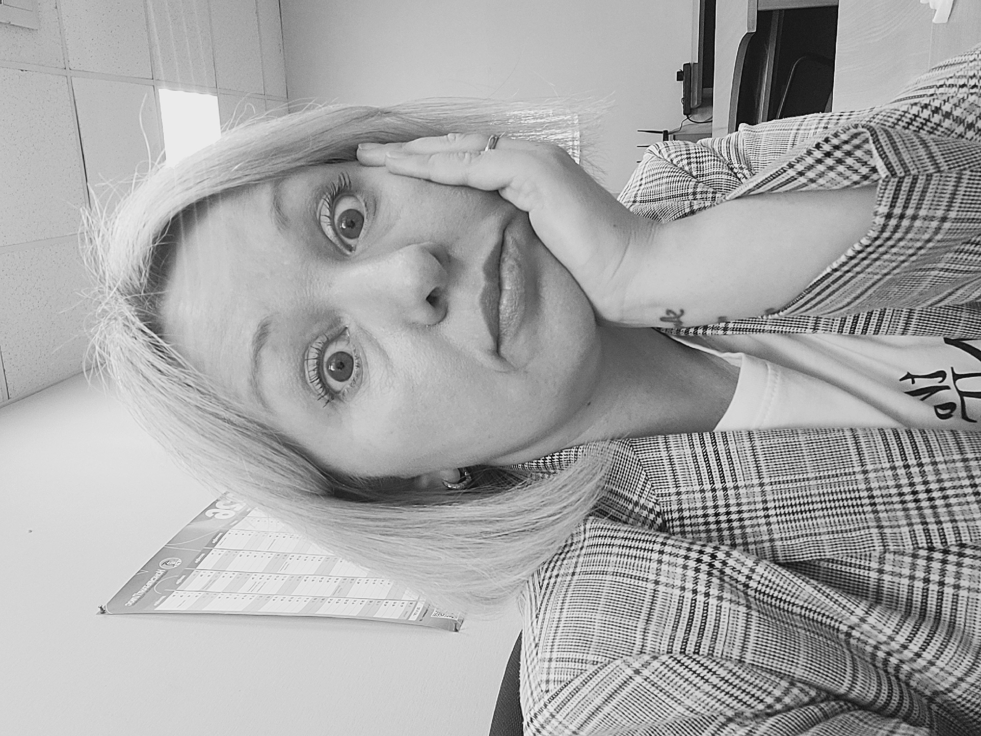A monochrome image shows a person with blond hair and light skin wearing a plaid blazer. The person's face is the primary focus, with a hand resting on their cheek. They have wide eyes and a slightly pouting expression. In the background, there is a calendar on the wall and a partial view of office furniture. There is no indication of a location within the city of Izhevsk, Russia.