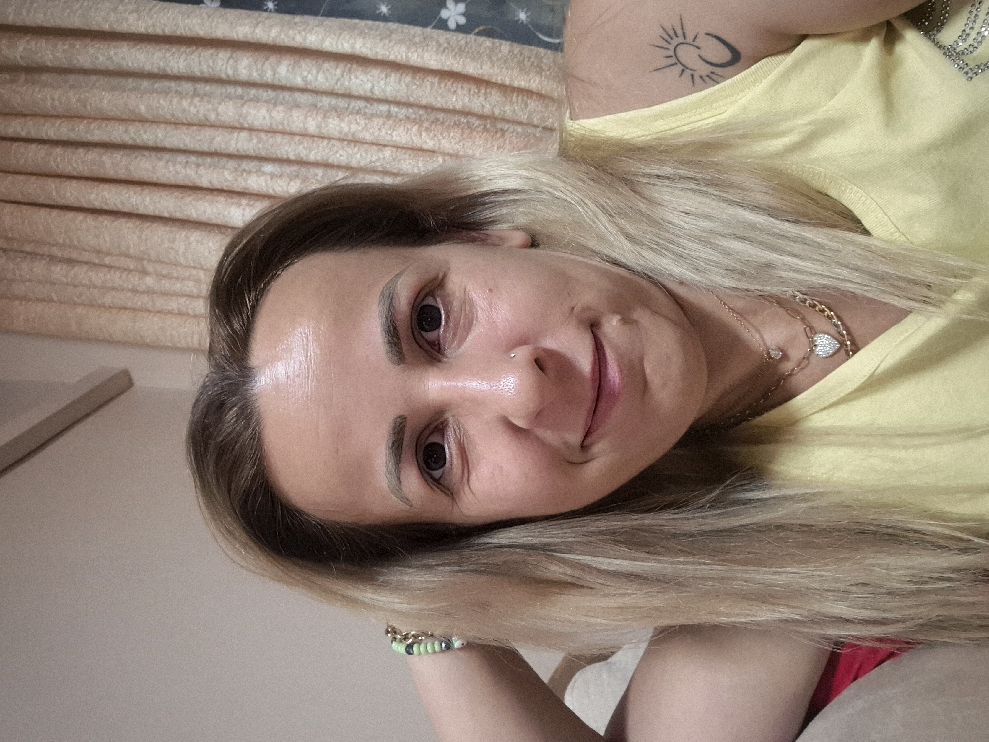 A woman is taking a selfie in what appears to be a home setting. She has blonde hair, light skin, and is wearing a yellow tank top. She is also wearing a necklace with a heart-shaped pendant and a bracelet on her left wrist. There is a tattoo on her right shoulder of a sun with rays and a crescent shape. In the background, there are curtains. The lighting suggests the photo was taken indoors, possibly during the day or with artificial lighting.