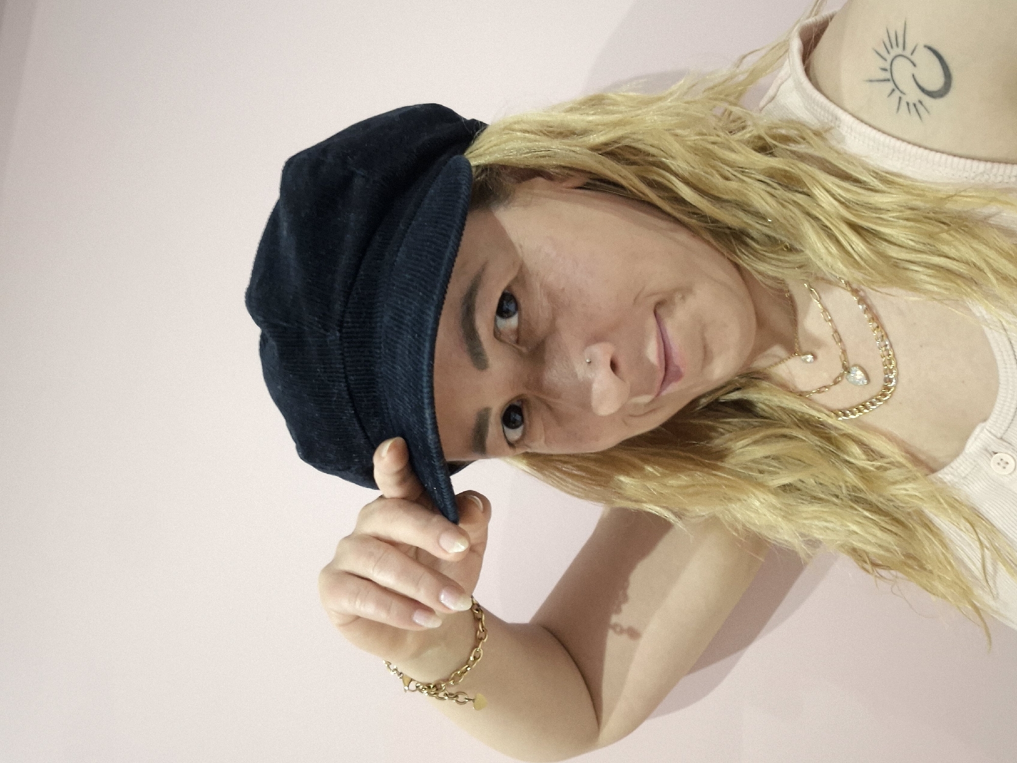 A woman is posing indoors, wearing a dark blue corduroy cap, a white button-down shirt, and gold jewelry. She has blonde hair and a nose piercing. A tattoo is visible on her upper chest. The background appears to be a light pink or peach-colored wall.