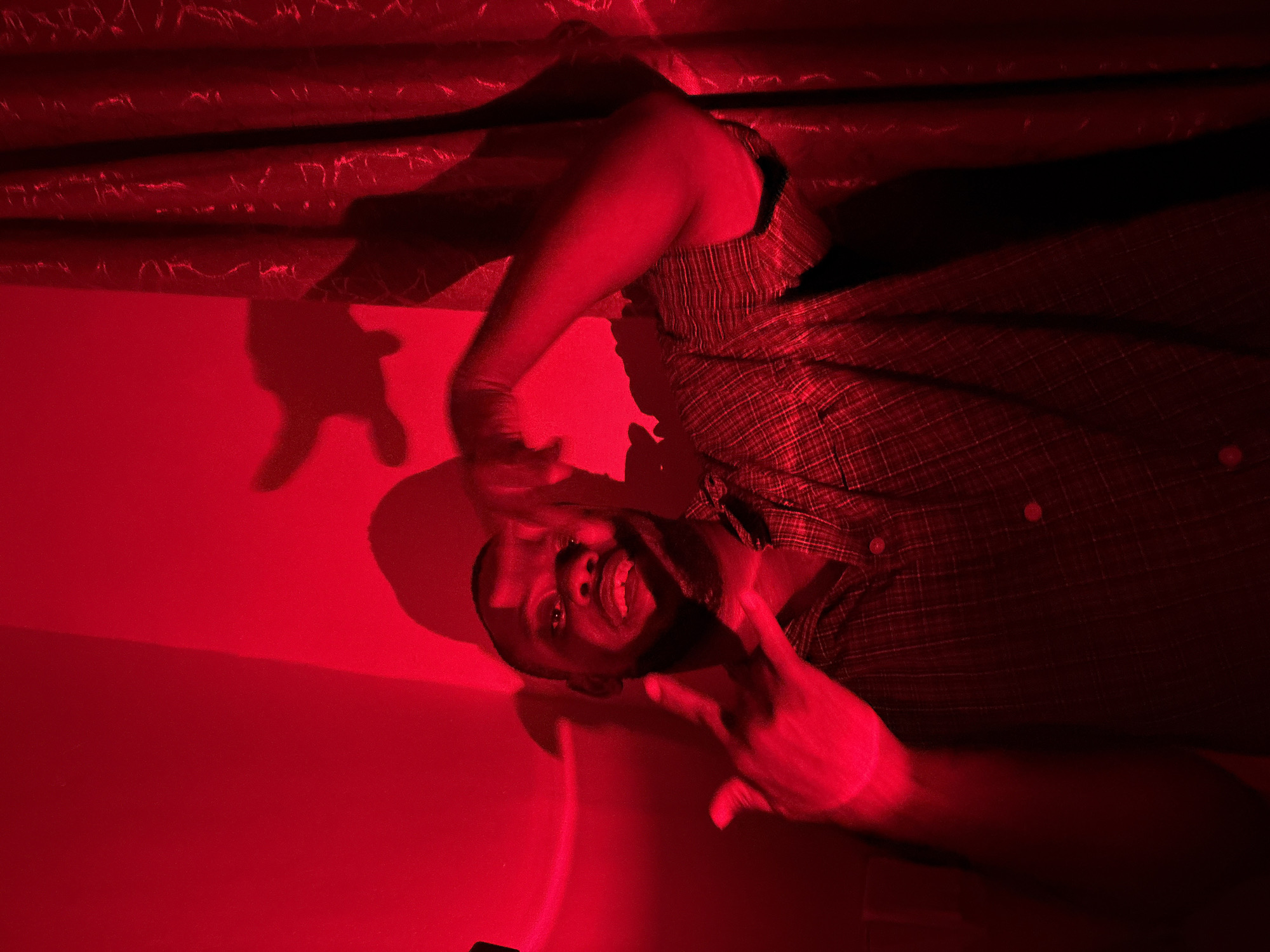 A man with dark skin is illuminated by intense red light. He is grinning and holding his hands up. He is wearing a button-down plaid shirt. Behind him is a wall, and above him is draped fabric or material. The scene is bathed in a single red light source. There are no specific identifiable details linking the image to Iddo Tudunwada, Nigeria.