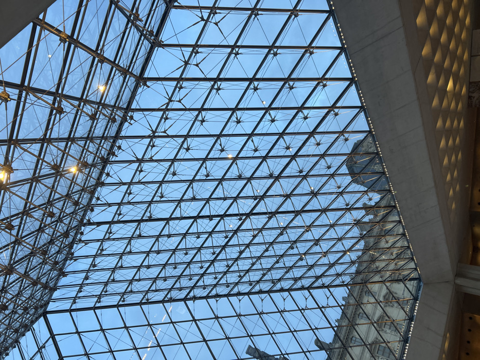 A geometric skylight is composed of numerous triangular glass panels held together by a black metal framework. The blue sky is visible through the glass. Architectural elements, including a turret-like structure, are visible through the glass. A concrete wall with a textured, diamond-patterned surface runs along the right side of the image. Some lighting fixtures are seen through the skylight.
