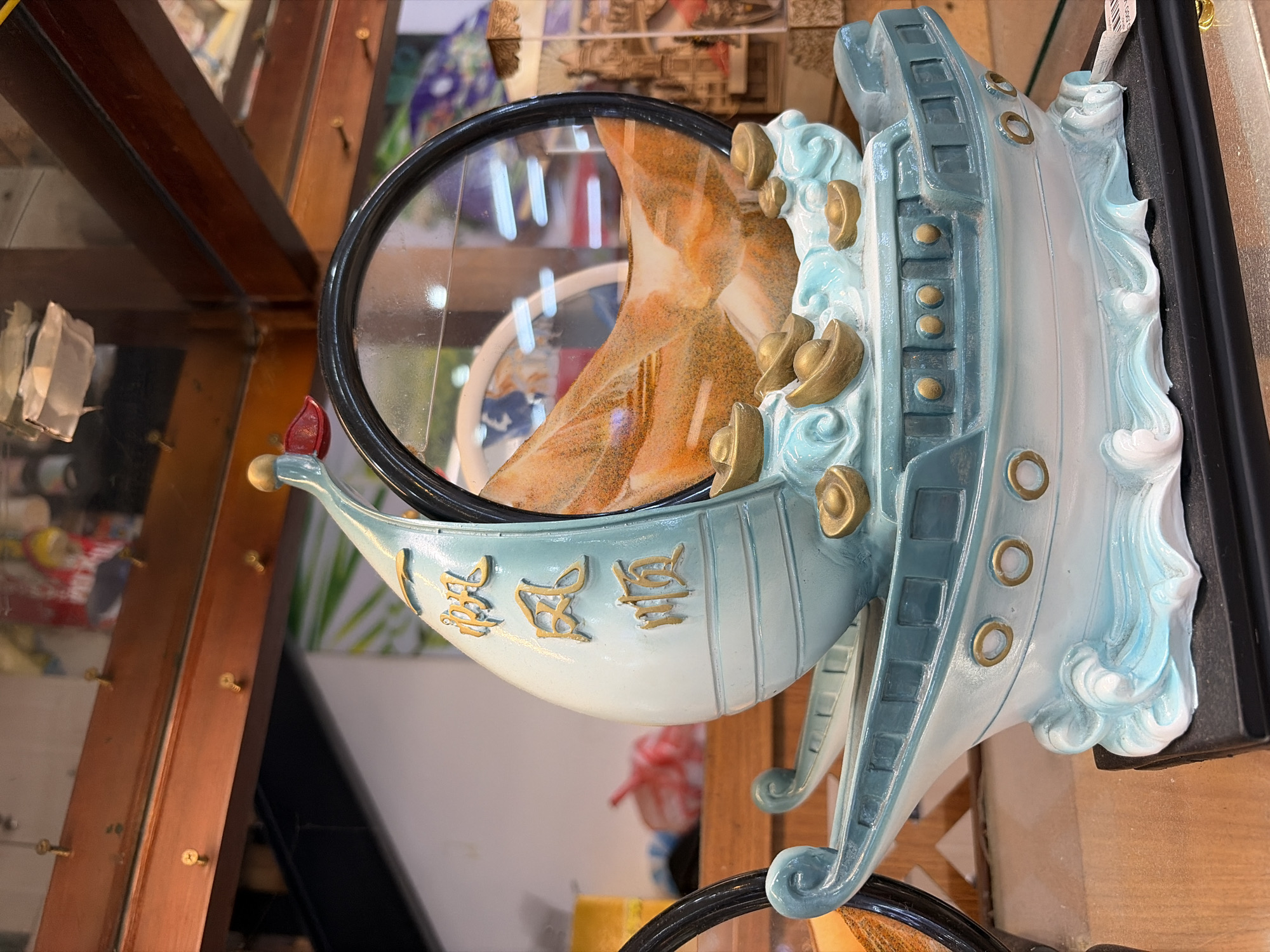 The image shows a decorative boat sculpture with Chinese characters inscribed on its sail, filled with orange sand. The sculpture features gold-colored ingots on top of stylized blue waves. The boat is placed on a black base. In the background, a wooden shelf with screws is visible, and behind that, there are blurry displays of other items and posters, suggesting an indoor retail environment. The overall scene is well-lit, with the focus on the detailed craftsmanship of the decorative boat. There are no people visible in the image, and the specific location in Đà Nẵng, Vietnam, cannot be determined from the visual information provided.