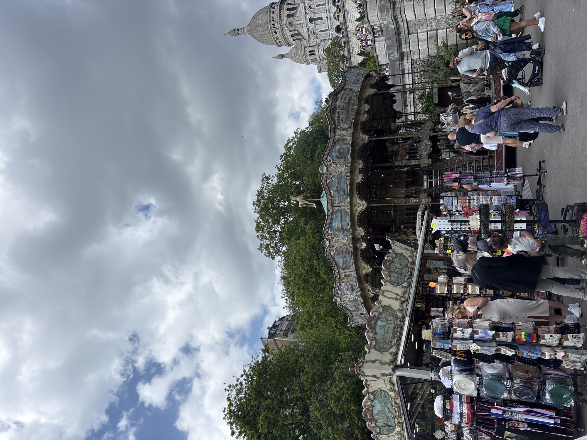 An eye-level shot captures a lively street scene in Montmartre, Paris, with the Sacré-Cœur Basilica dominating the background under a partly cloudy sky. The iconic white dome and towers of the basilica are visible, with numerous people gathered on its steps and surrounding area.
In the middle ground, an ornate carousel stands, featuring horses and other classic carousel figures. Its detailed facade shows decorative panels. A single white carousel horse is prominent on the left side of the carousel. Lush green trees line the area between the carousel and the basilica, and also extend towards the foreground.
The foreground is bustling with people and market stalls. Shoppers browse a variety of merchandise displayed on racks and shelves, including what appear to be textiles, hats, and souvenirs. Many individuals, adults and children, are walking, standing, or interacting with the vendors. One person is seen pushing a "BENTI" branded stroller. Visible text on the carousel includes "CARROUSEL DU TER...", likely referring to the "Carrousel du Tertre." On one of the market stalls, the word "CHOU" is visible on a box.
The weather appears to be overcast with some patches of blue sky peeking through, suggesting a partly cloudy day. The overall impression is one of a busy tourist area, typical of Montmartre, with leisure activities and commercial offerings.