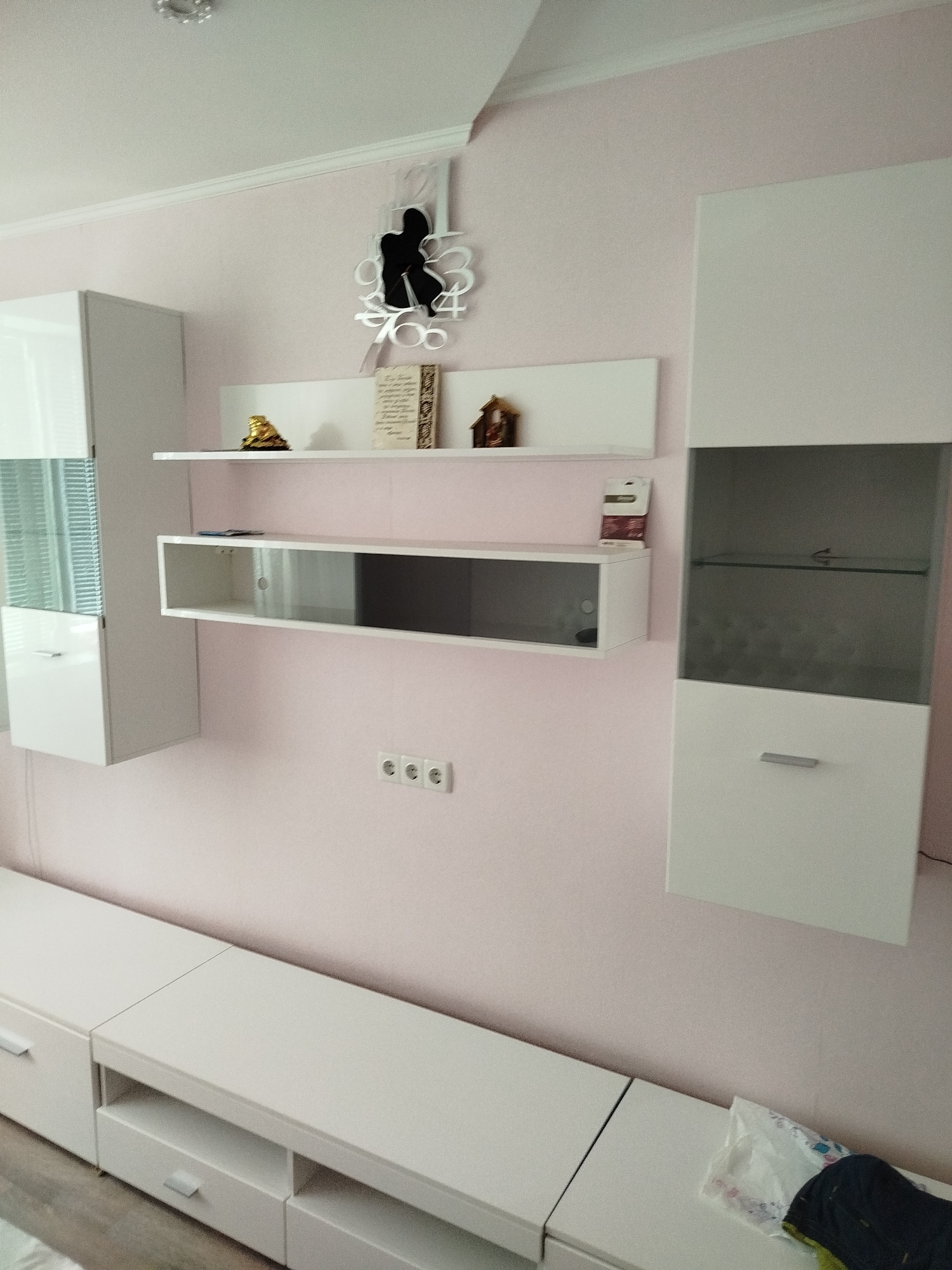 The image shows a modern living room with white furniture against a pale pink wall. On the wall, there are several white shelves, a decorative clock shaped like a person, and three electrical outlets. The shelves hold a few decorative items, including a small golden statue, a framed piece of text, and a miniature house. To the left and right of the shelves are tall white cabinets, one with a glass-fronted section. Below the shelves, a long, low white media console with drawers and open compartments spans the width of the wall. The floor appears to be wooden, and a white plastic bag is visible on the media console.