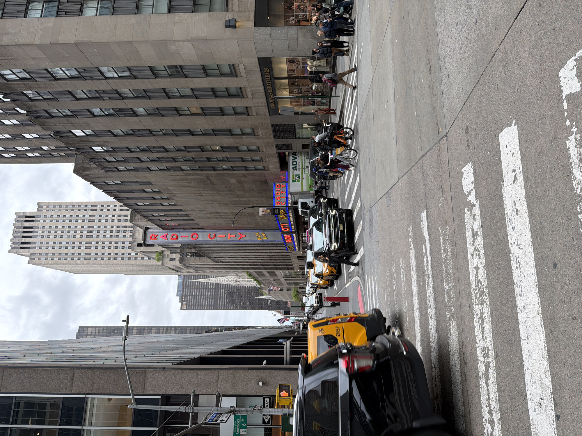 This image depicts a bustling daytime street scene in a major city, identifiable as New York City due to prominent landmarks and street signs. The perspective is from the sidewalk, looking down a wide street with multiple lanes of traffic and tall buildings on both sides. The sky is overcast, suggesting a cloudy day.
Numerous people are visible, engaged in typical urban activities. Several pedestrians are on the sidewalk, with a crowd gathered outside a storefront. One person, wearing a brown jacket and carrying a backpack, is midway across a white-striped crosswalk.
The street is filled with various vehicles, including several yellow New York taxis, black cars, and a white commercial truck. Some bicycles, possibly rental bikes, are also seen parked or being ridden near the crosswalk. Traffic appears to be moving or waiting, indicating a busy period.
Prominent architectural features include modern high-rise buildings and older concrete structures with many windows. A large, illuminated neon sign for "RADIO CITY MUSIC HALL" stands out on one of the buildings, adding a famous landmark to the scene. Another notable storefront sign reads "BROOKS BROTHERS." A street sign clearly indicates "Americas 6 Av," confirming the location. Other visible text includes "ONE WAY" on a traffic sign, "ADVA Moving You" on a truck, and "2N57" on a yellow vehicle. The overall impression is one of a lively, active urban environment.