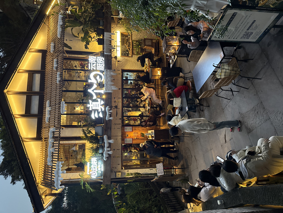 The image depicts a multi-level restaurant located in Nan'an District, China, specifically identified by the illuminated signage as "滇个勺 云南厨房" (Dian Ge Shao Yunnan Chufang, or Yunnan Kitchen). The establishment is observed at dusk or evening, with prominent warm artificial lighting illuminating its facade and outdoor dining areas.
The building is a two-story structure with a dark roof and light-colored walls, featuring large glass windows on the upper level. Decorative white elements resembling melting snow or icing are affixed along the roofline and above the lower windows. Additional signage reads "心动榜 上榜餐厅 2023" (Heartbeat List, Listed Restaurant 2023). Potted plants are visible both inside and outside the building, enhancing a lush aesthetic.
The scene includes approximately two dozen individuals, predominantly adults, engaged in dining and social activities.
* **Upper Level (Indoor):** Multiple groups are seated at tables visible through the large windows, appearing to dine or socialize in an illuminated interior.
* **Lower Level (Outdoor/Patio):** This area functions as an outdoor dining and waiting space on a stone-paved surface.
* To the left, a group of approximately eight individuals is either waiting or socializing. Some are seated on a low stone wall or bench, while others stand. Several are looking at mobile phones. A wooden railing and staircase descend further to the left, with one individual visible on the stairs.
* Near the main entrance, two individuals appear to be interacting with staff or reviewing information, standing beside a small service counter or podium.
* In the central and right portions of the patio, several groups are seated at various tables (square, rectangular, round) with chairs, actively dining or conversing. Some tables are currently unoccupied.
* Two freestanding, transparent, rectangular outdoor heaters with visible flames provide warmth and ambiance in the dining area.
* A large sign with text and a QR code is visible on the right, partially obscured by foliage.
The overall environment suggests a popular, vibrant dining establishment operating in the evening.