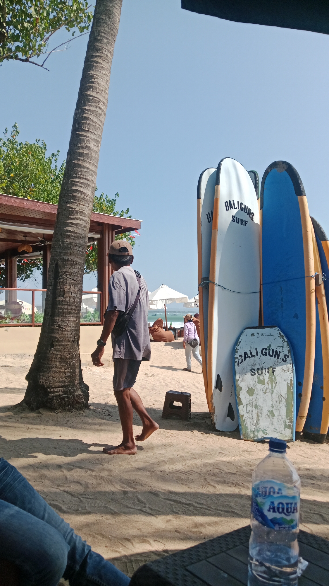 On a sandy beach in Kuta, Indonesia, a person wearing a grey t-shirt, grey shorts, and a cap walks past a large palm tree. A row of surfboards stands upright to the right, some bearing the inscription "BALIGUNS SURF." In the background, white beach umbrellas and beanbag chairs suggest a relaxed setting. A bottle of Aqua water sits on a table in the foreground. The sky is clear blue.