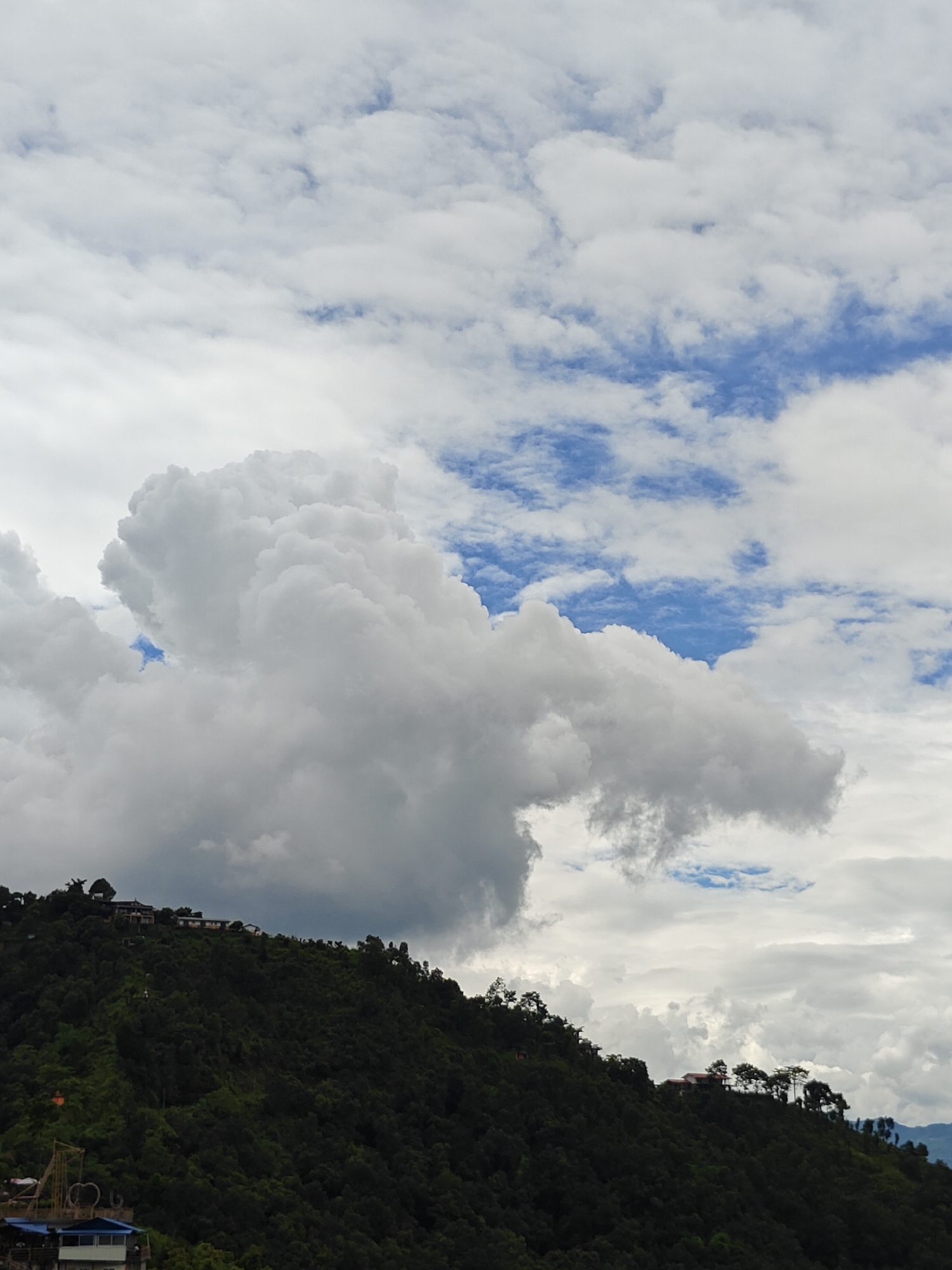 The image captures a panoramic view of the lush green hills of Pokhara, Nepal, under a dramatic sky filled with white, fluffy clouds interspersed with patches of blue. The foreground is dominated by the steep, densely forested slopes of a hill. Several buildings and structures are visible nestled on the ridgeline and upper slopes of the hill, suggesting a populated area or resort on the elevation. Trees, varying in height and density, cover the hillside, creating a textured, dark green landscape. In the lower left corner, parts of man-made structures, including a building with a blue roof and some sort of yellow metal framework, are visible, hinting at the edge of a developed area or a viewpoint. The sky above is a spectacle of dynamic cloud formations, with large, voluminous clouds creating shadows and highlights against the brighter sky. The overall impression is one of natural beauty combined with human habitation on a mountainous terrain.