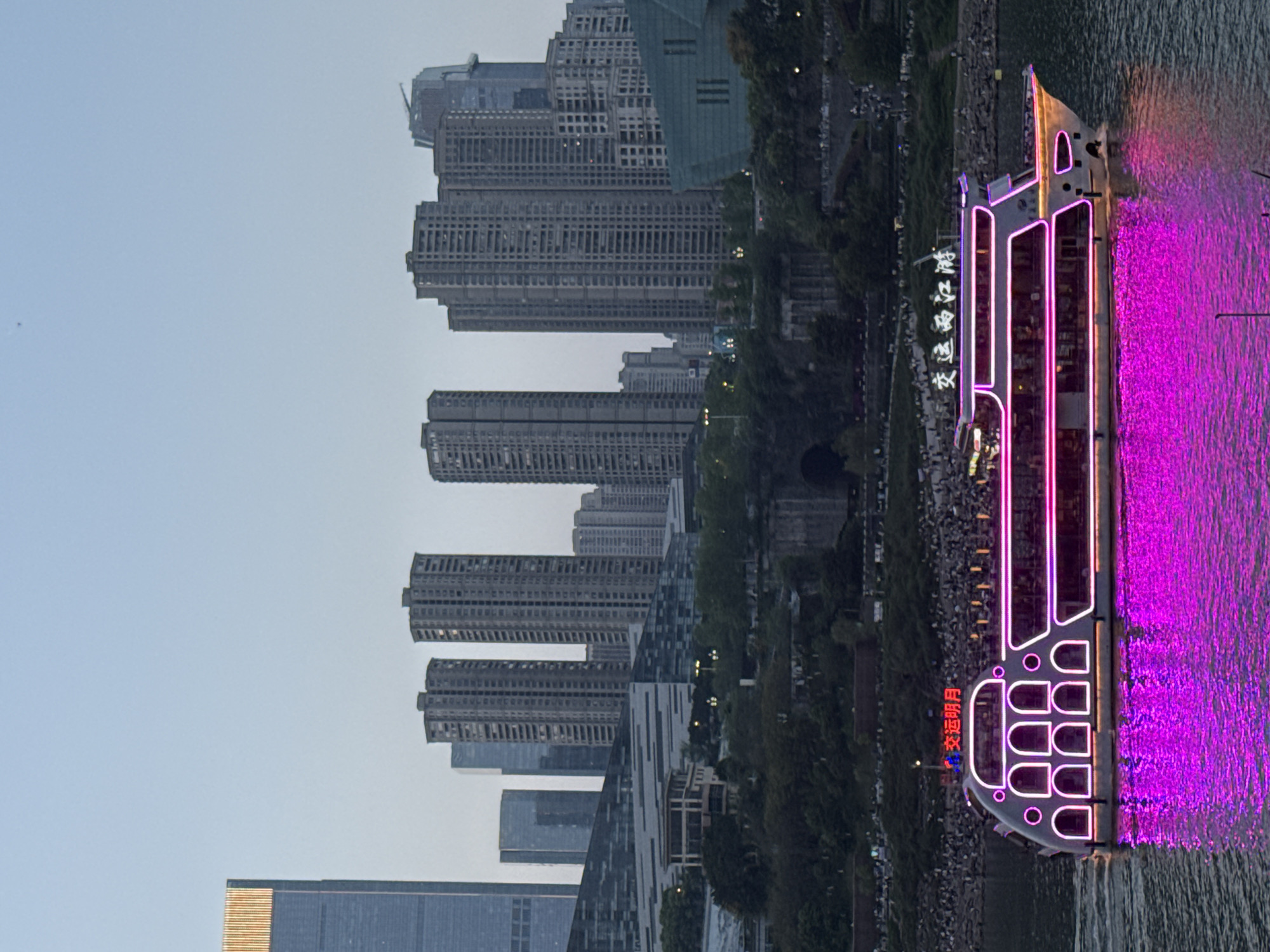 The image shows a cityscape, with a river in the foreground and a line of modern high-rise buildings in the background. On the right side of the river is a boat illuminated with bright pink lights. Along the shoreline, there appear to be trees and a gathering of people. The location is Yuzhong District, China.