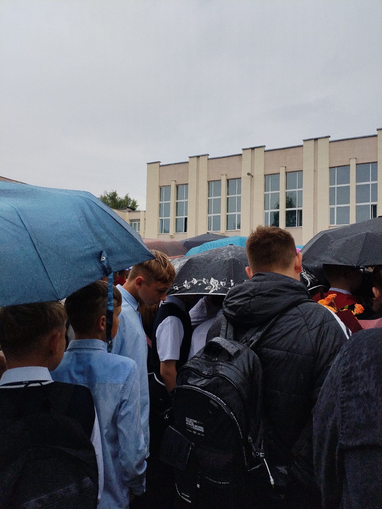 In Bialynichy, Belarus, a crowd of people, primarily young males, stand under umbrellas on an overcast day. The setting appears to be outside a building with a pale facade and numerous rectangular windows, possibly a school. Several individuals are wearing light blue shirts, some with dark vests or jackets. Backpacks are visible, including a black one with white text. The ground is wet, indicating rain. Some individuals are holding bouquets of orange flowers. The atmosphere is somewhat somber due to the weather and the overcast sky.