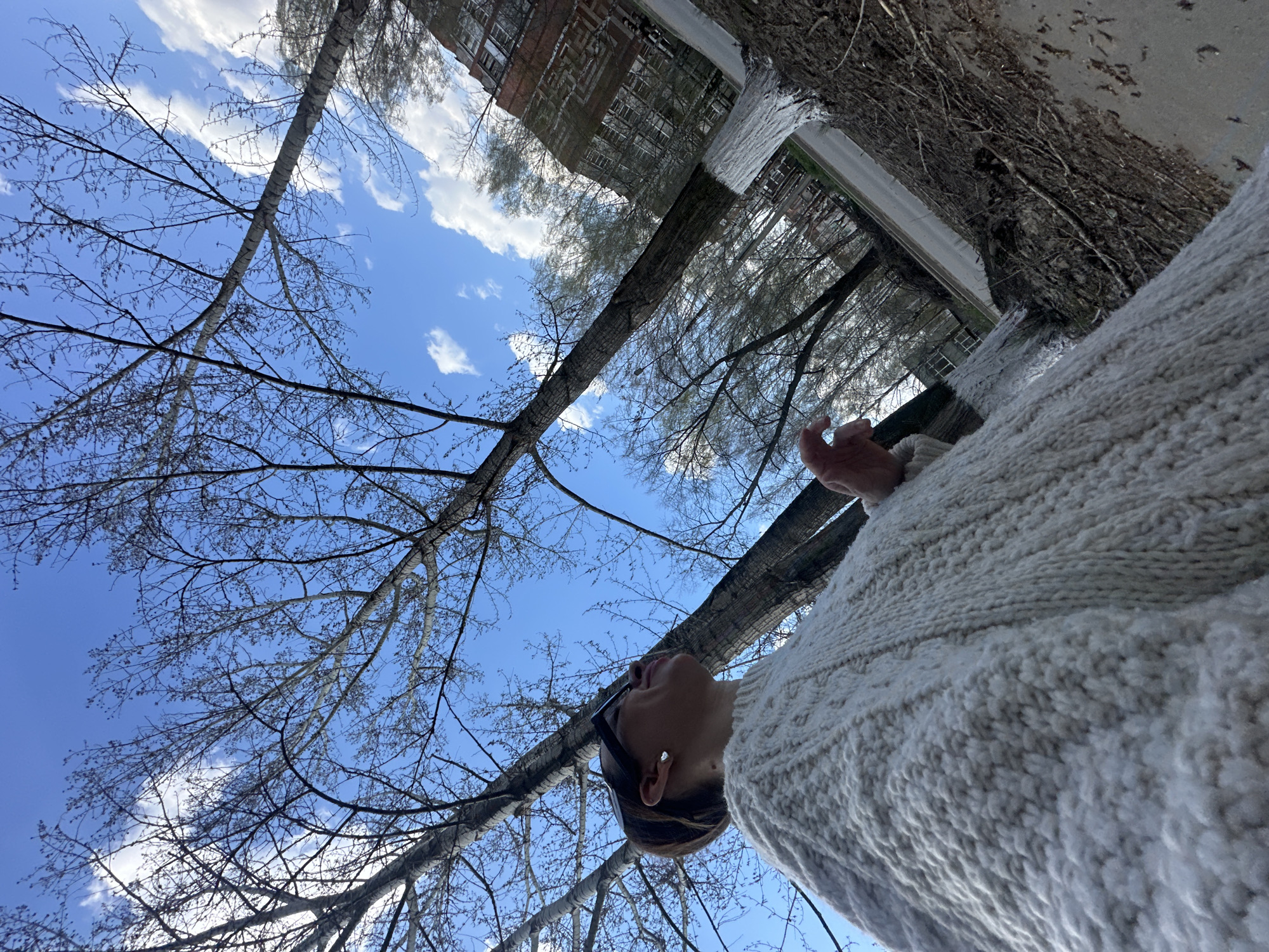 A low-angle shot depicting a woman in a white sweater looking up at bare trees against a bright blue sky with scattered white clouds. The branches are sparse, indicating either winter or early spring. The trunk of a tree runs diagonally across the center of the frame. In the background, a brick building is visible, which suggests an urban setting. The location is Votkinsk, Russia. The woman's face is partially visible, showing her chin, nose, and a hint of a smile. She appears to be standing on a paved surface. There are architectural details in the background, particularly parts of building facades. The environment is a public outdoor space.