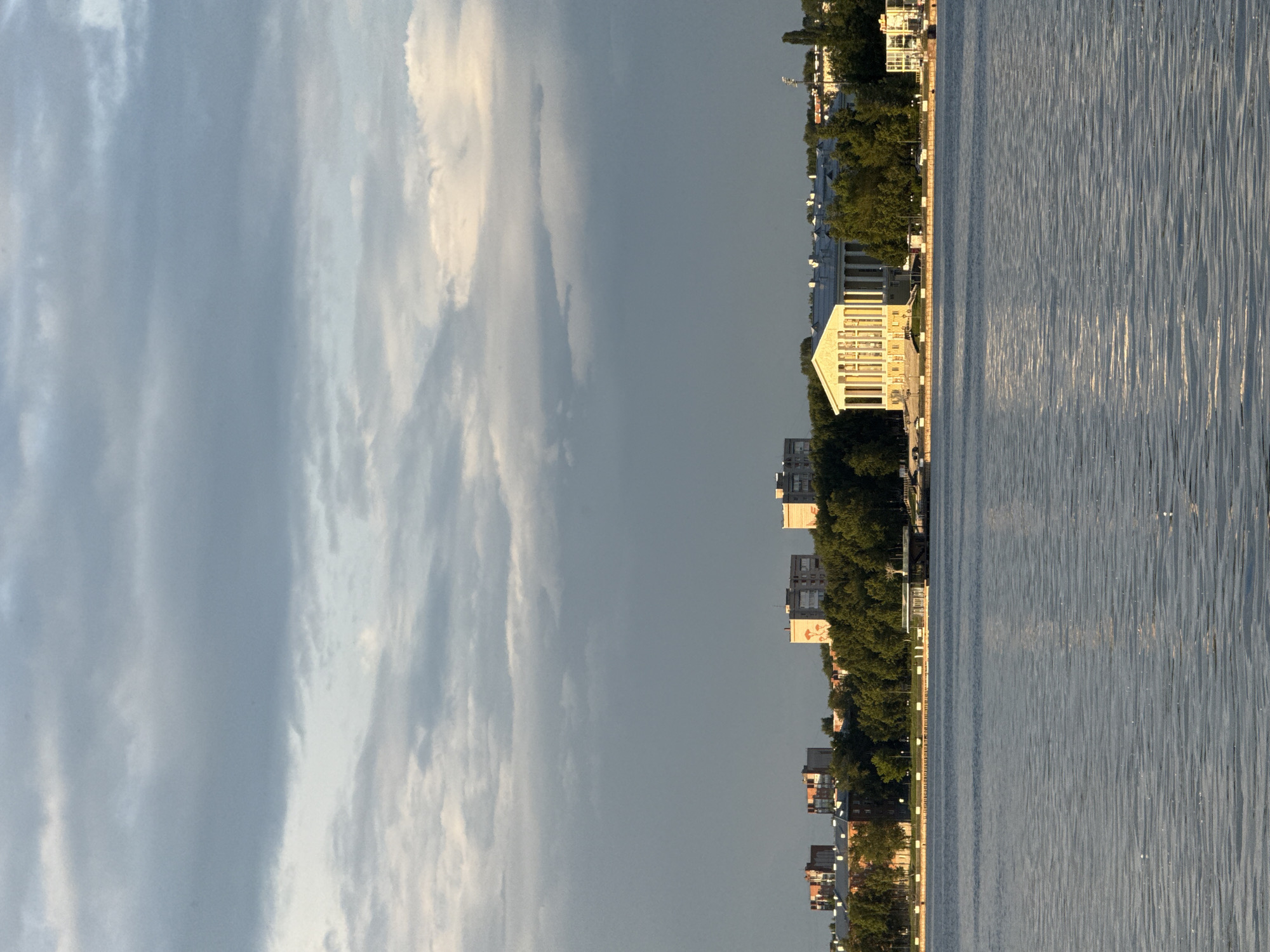 The image captures a wide shot of a calm river in Votkinsk, Russia, reflecting the overcast sky and the cityscape on the opposite bank. Dominating the middle ground is a grand, Neoclassical building with a portico of numerous white columns, illuminated by the golden light of a setting sun. This building is likely the Votkinsk State Geological Museum or the House of Culture, a prominent landmark in the city. Behind it, a dense line of green trees separates the structure from a series of apartment buildings in varying architectural styles. The buildings on the far left appear older, constructed of brick, while those towards the right are more modern. The riverfront is lined with a promenade and some smaller structures, including a covered pavilion. The water's surface is rippled, indicating a gentle breeze. The sky is a mosaic of grey and white clouds, with patches of brighter light suggesting the sun is breaking through. There are no visible people in the frame.