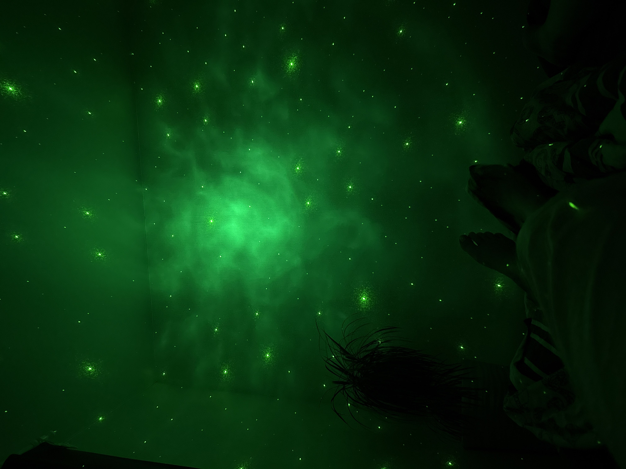 This media file captures a dimly lit room adorned with a green galaxy projector, casting a starry, nebulous glow on the walls and ceiling. The ambiance suggests a nighttime setting, possibly within a home or a dedicated relaxation space. In the right portion of the frame, a person's legs and feet are visible, positioned as if they are reclining or lying down. Their attire appears casual, with patterned fabric peeking through the low light. To the left of the frame, a dark, wispy object with elongated strands extends into the illuminated space, its nature unclear in the subdued lighting, possibly a piece of furniture or an artistic element. The scene evokes a sense of calm and wonder, as if one is immersed in a cosmic landscape. No visible text is present in the image. The location context of Perm, Russia, is noted but not visually evident within the frame itself.