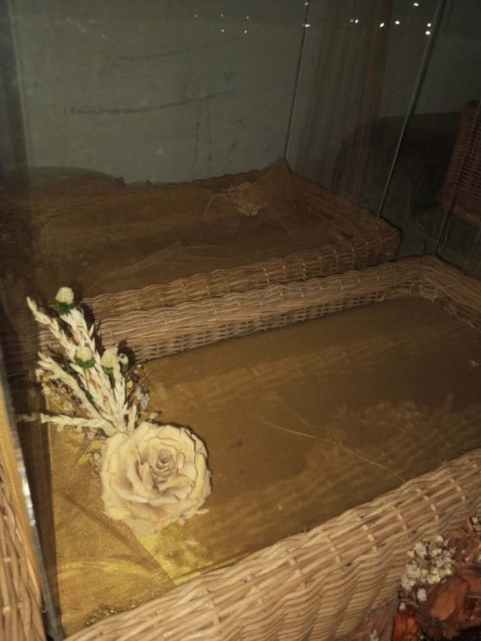 Two brown woven baskets with gold fabric are displayed in a glass case. One basket is slightly behind and above the other, partially covered by a thin, textured brown cloth. The visible basket features a large, artificial rose and assorted small, dried flowers in its corner. The background is a muted green, reflecting light and adding to the display's depth. The basket weave is tight and uniform.