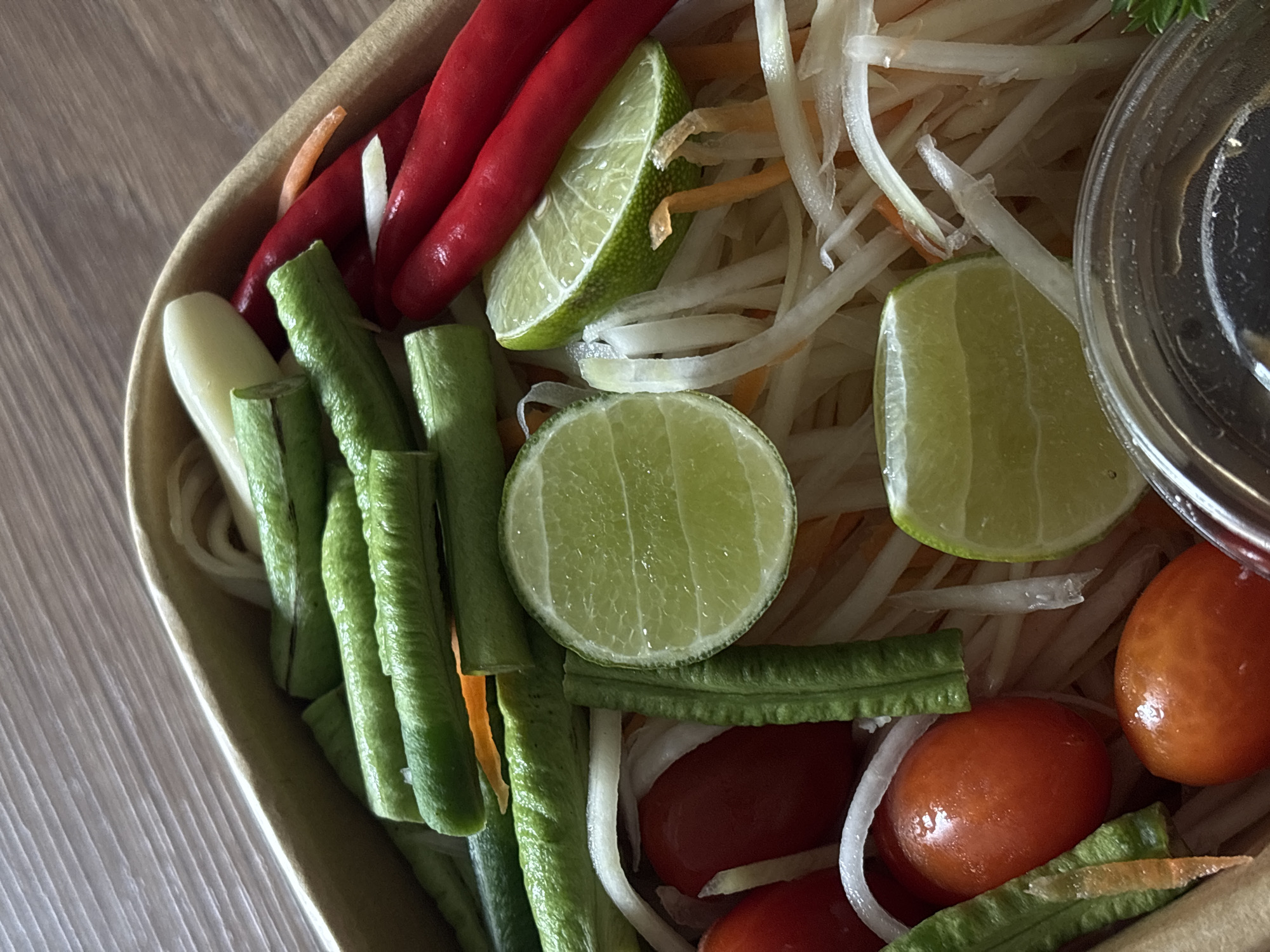 The image presents a vibrant, close-up view of a fresh Thai salad, consistent with Som Tum (papaya salad), served in a light brown, possibly disposable or eco-friendly, container. The dish is packed with a colorful array of ingredients.
Dominating the salad are numerous thin, white shredded strips, likely green papaya, mixed with finer orange shreds, presumably carrots. Several bright red, long chili peppers are arranged prominently towards the top left, suggesting a spicy flavor. Distributed throughout the salad are multiple slices and wedges of fresh green lime, indicating a citrusy element.
Also visible are segments of vivid green long beans, cut into pieces, and small, round, reddish-orange tomatoes, similar to cherry tomatoes, concentrated towards the bottom right. Thin white rings, possibly sliced onions, are interspersed among the tomatoes and other ingredients. A small, clear plastic cup, likely holding a dressing or sauce, is visible in the upper right corner of the container.
The container rests on a light brown wooden surface with a distinct grain pattern. The overall composition highlights the fresh and colorful ingredients of a classic Thai dish, aligning with the location context of Choeng Thale, Thailand. No visible text is present in the image.