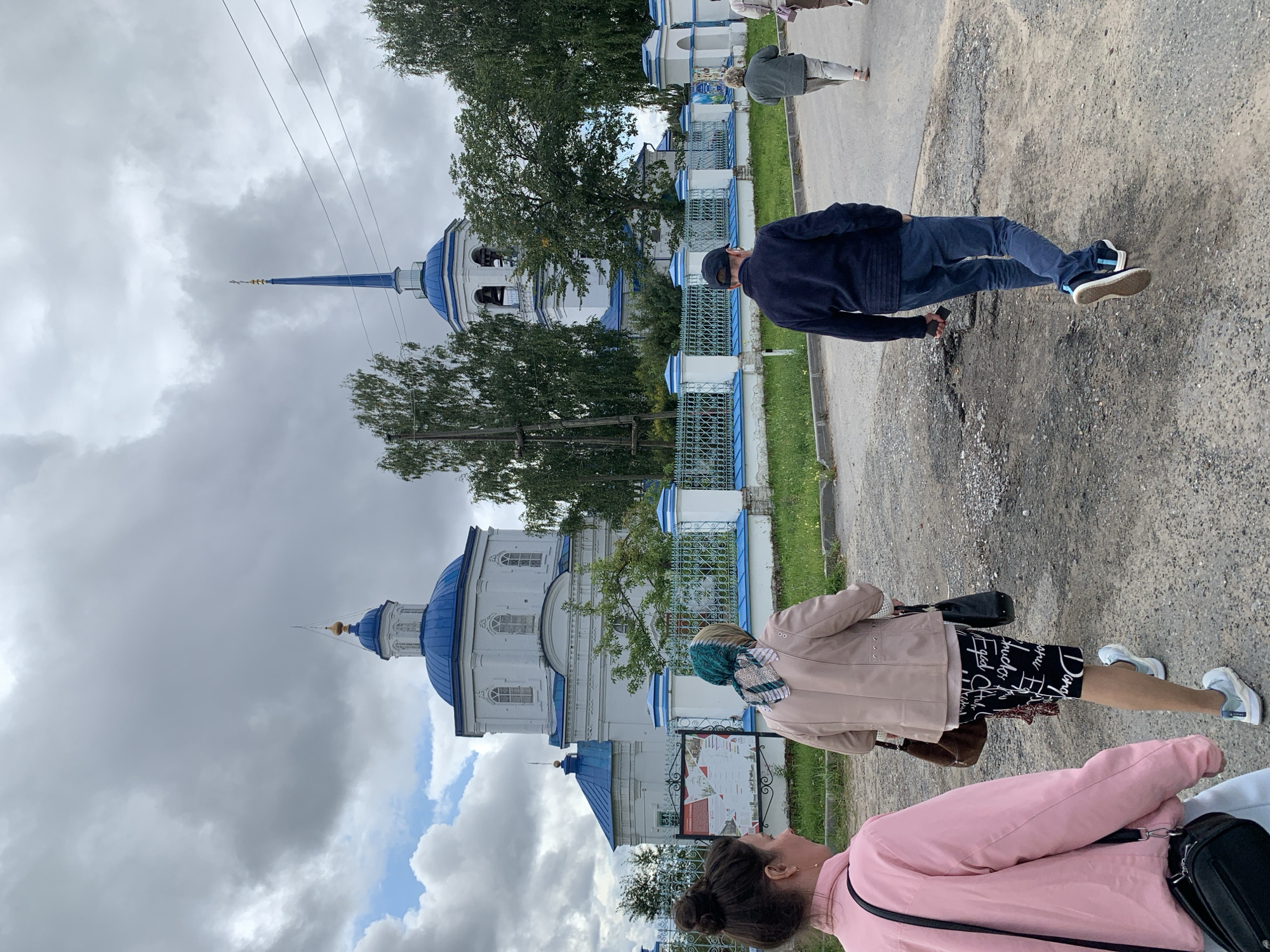 A group of four individuals are walking on a gravel road towards a white church with blue domes and a blue spire. The church appears to be the Holy Trinity Cathedral, located in the city of Orel, Russia. The sky is overcast with gray clouds. The people are walking away from the camera, and their faces are not visible. The person in the foreground on the left is wearing a pink jacket and a black crossbody bag. To their right, a person in a light-colored jacket and patterned skirt is walking. Further ahead, a man in a dark blue sweater and blue pants is walking, and behind him, a person in a gray jacket and light-colored pants is walking. A green metal fence with white posts and blue trim runs along the side of the road in front of the church. Trees are visible behind the fence and around the church.