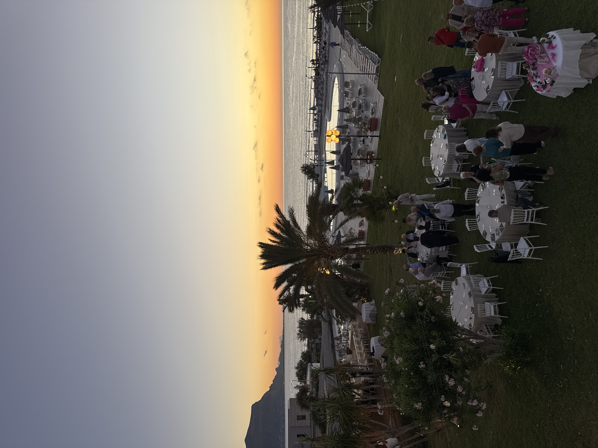 The image depicts an outdoor evening event set against a coastal landscape at sunset. The location is identified as Isola delle Femmine, Italy, with a view of the sea and a prominent mountainous coastline in the distance. In the foreground, a grassy lawn is set up with several round tables draped in light-colored tablecloths, surrounded by white chairs. Guests, appearing to be adults, are mingling and seated at some of the tables. One table in the lower right is decorated with pink floral arrangements. Palm trees and other vegetation are scattered throughout the scene. In the mid-ground, a swimming pool area is visible with lounge chairs and umbrellas. The sky displays a gradient of colors from pale blue at the top to warm orange and yellow hues near the horizon, indicating sunset. The overall atmosphere suggests a social gathering, possibly a wedding reception or a special event, taking place at a resort or hotel by the sea.