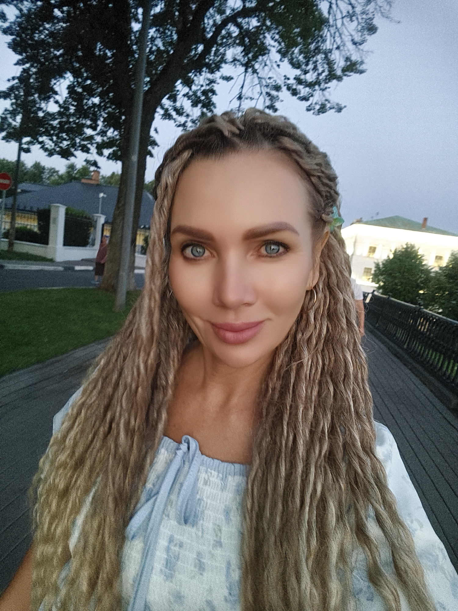 A woman with blonde dreadlocks is taking a selfie outdoors. She has blue eyes and is wearing a light blue and white patterned top. Behind her, a tree, a street, and a building can be seen. The location is not specified in Yaroslavl, Russia.