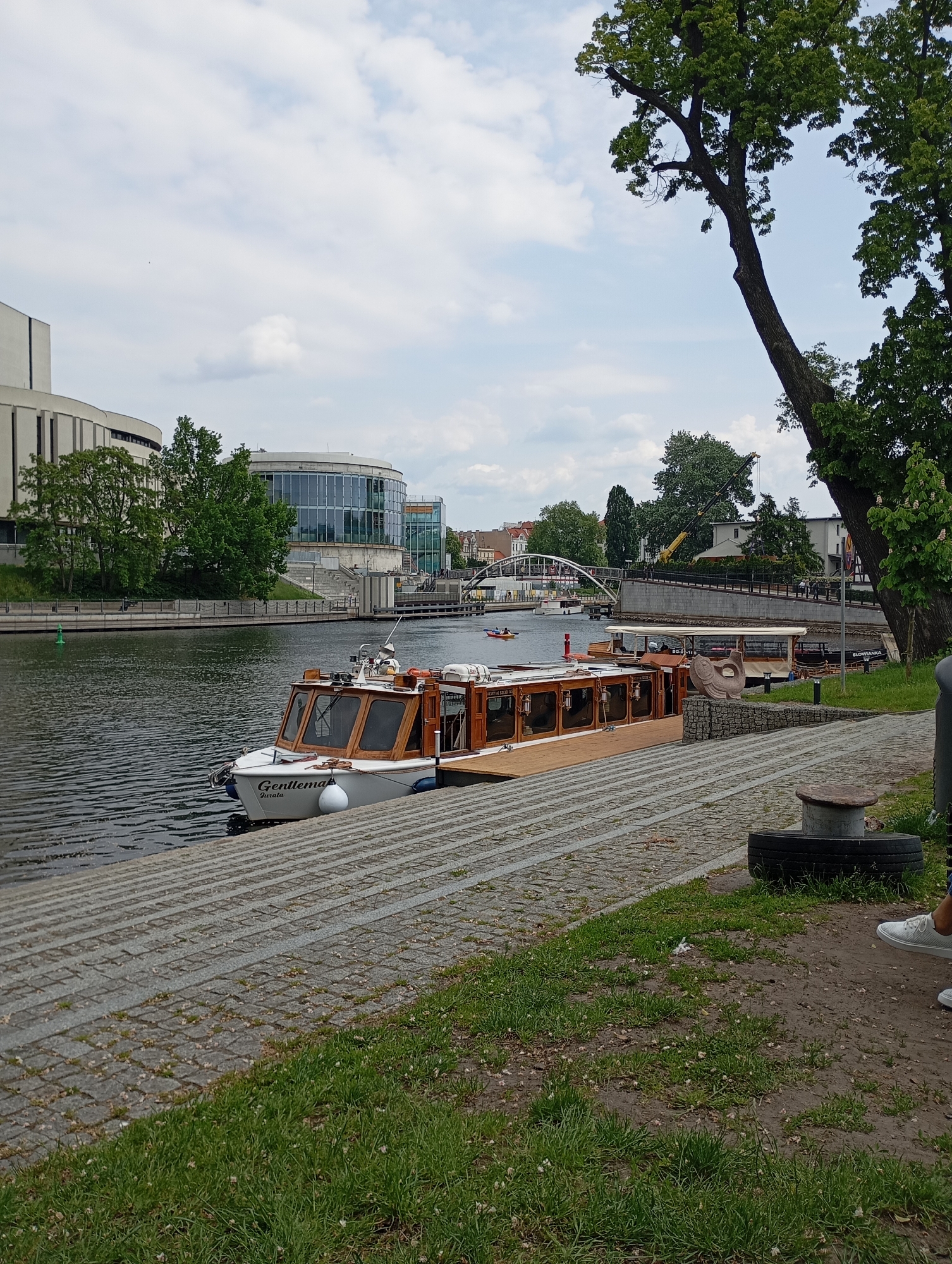 The image shows a view of the Brda River in Bydgoszcz, Poland, on a partly cloudy day. A tourist boat named "Gentlemar" is docked on the riverbank. Other docked boats are visible in the background. The Opera Nova building is partially visible on the left. A white arched bridge is in the background. A tree is on the right side of the frame. Part of a person wearing white shoes is on the right, possibly sitting down. The riverbank is lined with stone paving.