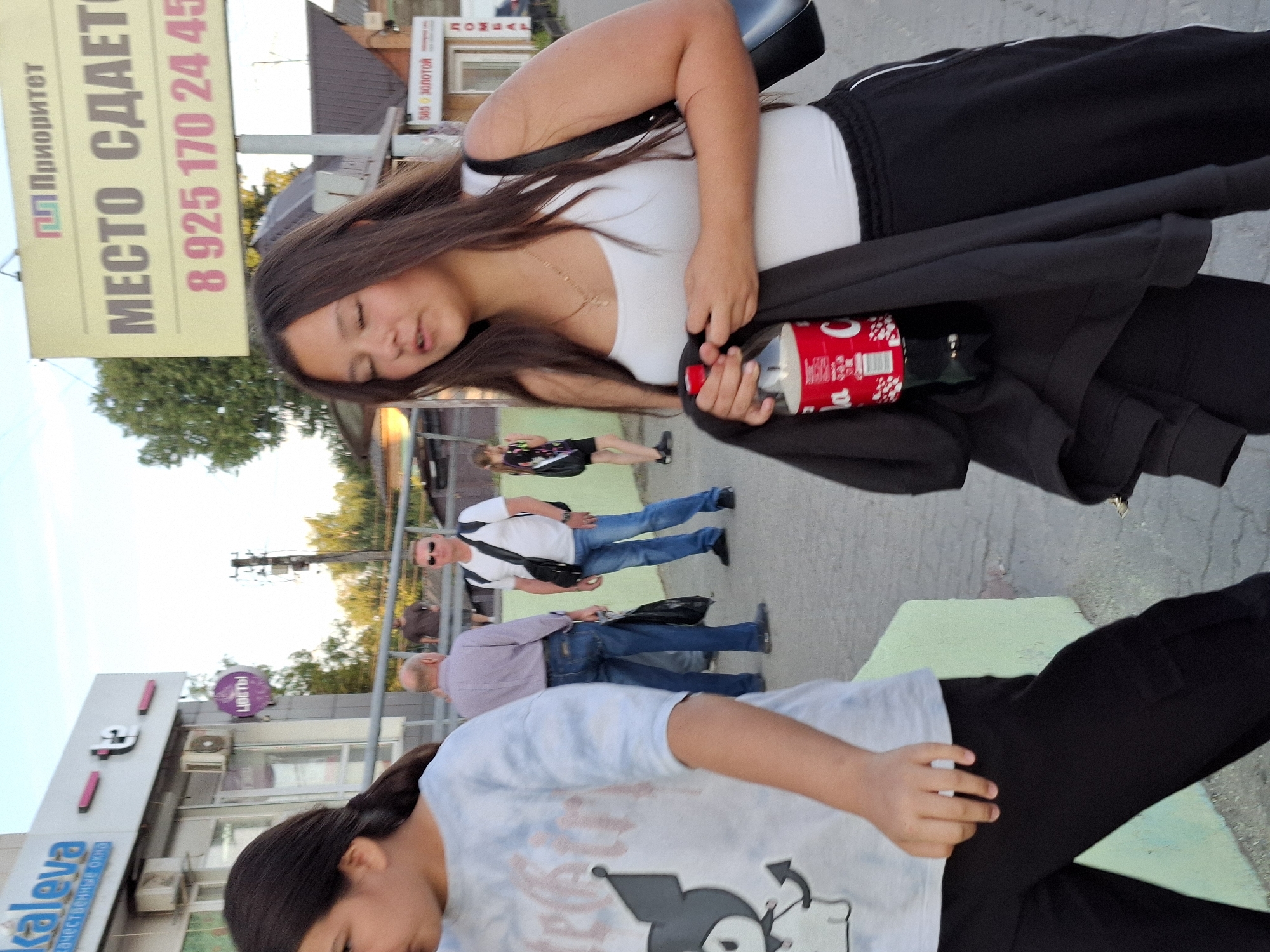 In an outdoor urban setting, a young girl in a light-colored t-shirt and black pants is walking, holding a bottle of soda and a dark jacket. To her left, another young girl in a graphic t-shirt and black pants is also walking. In the background, two men are standing on a sidewalk. One man wears a purple shirt and jeans, while the other wears a white t-shirt, jeans, and sunglasses, with a bag slung over his shoulder. A yellow billboard with Russian text is visible in the upper right portion of the image. The environment includes buildings with signage, trees, and a cloudy sky.