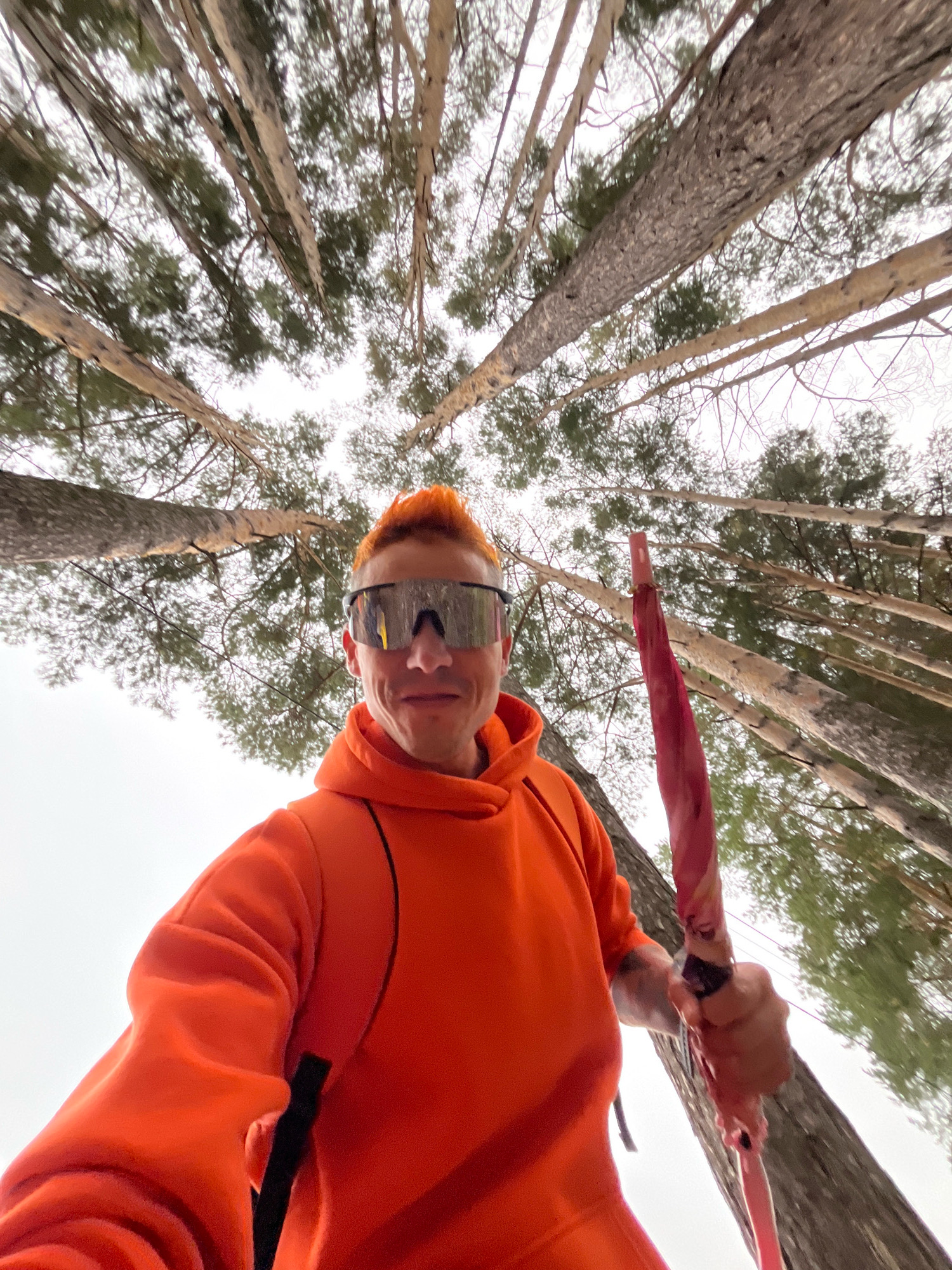 In the city of Chaikovsky, Russia, a person with bright orange, spiked hair and reflective sunglasses is taking a selfie outdoors. They are wearing an orange hoodie and holding a red object that appears to be a pink umbrella. The person is positioned in a forest with tall trees extending upwards towards a cloudy sky. The camera angle is from below, looking up at the person and the surrounding trees.